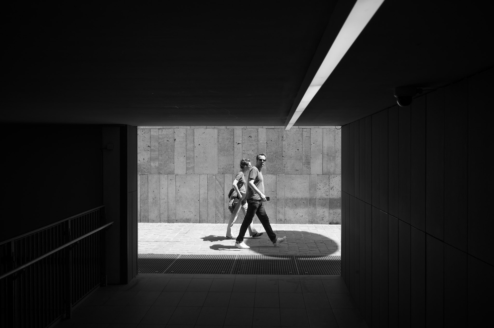 captured in munich's city center, this photograph perfectly encapsulates a fleeting moment in the urban landscape. two pedestrians are seen mid-stride as they exit an underpass, the contrasting light and shadow creating a striking frame around them. the architectural lines and the simplicity of the scene draw attention to the subtle interactions and movements within the city. this black-and-white image highlights the elegance of everyday life and the beauty found in spontaneous, unplanned moments.