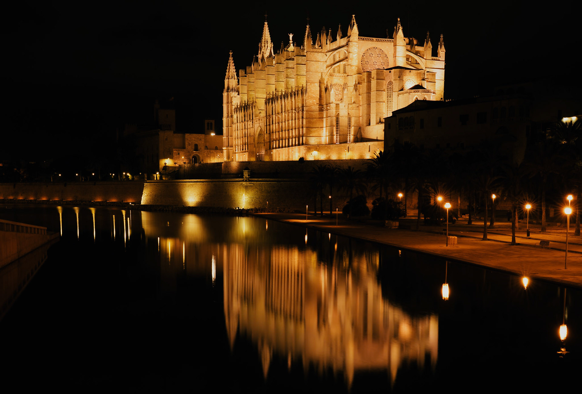 palma's cathedral, known as la seu, stands as a beacon of gothic architecture by night. its sandstone walls and flying buttresses are illuminated, casting a golden glow over the still waters. this majestic structure reflects not just on the surface of the water, but in the heart of mallorca's history, culture, and spirituality. built on the site of a former mosque, the cathedral's construction began in the 13th century and continued for centuries, allowing a blend of styles and the hands of many architects, including the famous antoni gaudí, to grace its form. at night, its presence is both haunting and serene, a watchful guardian over the city's slumbering streets.