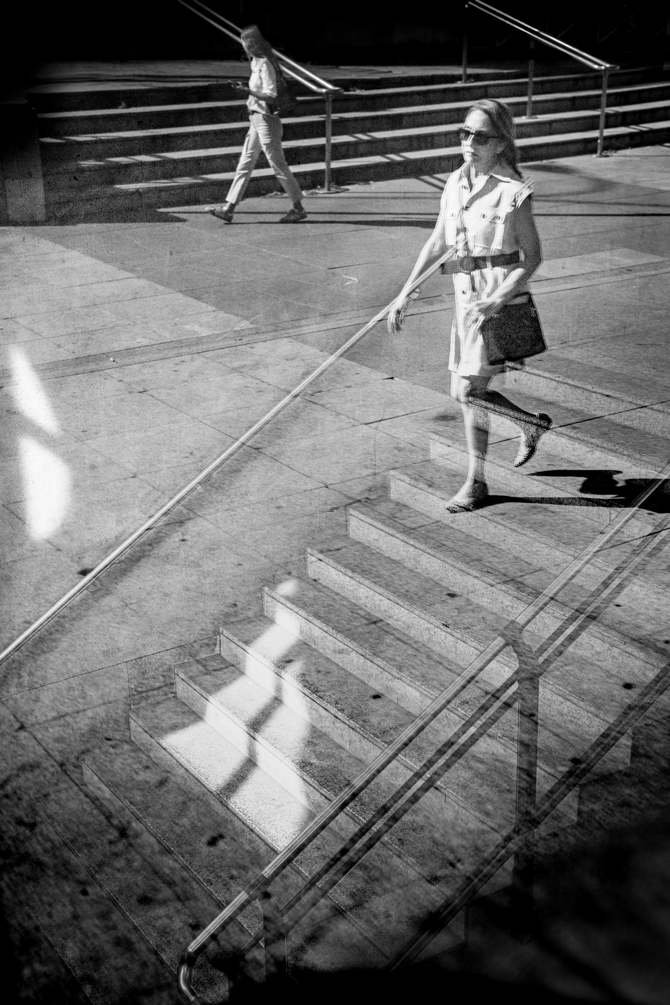 captured at madrid's "nuevos ministerios" train station, this photo was taken from outside on a window ledge, angled to photograph the staircase within, while incorporating the reflection of the exterior. it creates an illusion where the woman appears to be walking down the stairs inside, although her shadow clearly shows she is not. the stairs in the background are also from the outside. i found it interesting how two similar geometric motifs (the two staircases) merge, blending the outside with the inside, creating a deceptive narrative.