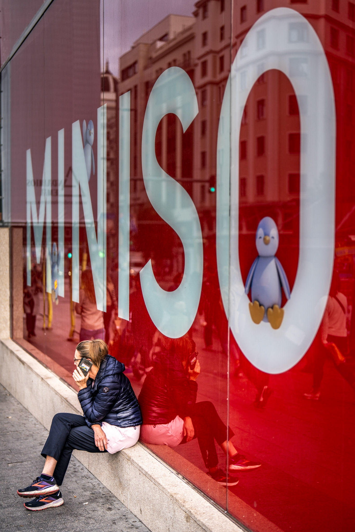 on madrid's gran via, moments pass and intertwine—layers of life against a vibrant cityscape. here, a lone woman finds a quiet space, her reflection mingling with passersby in the bustling reflection of a bright storefront. her thoughtful posture against the vivid red backdrop becomes a slice of solitude amidst the noise, her reflection an echo of her own moment.
