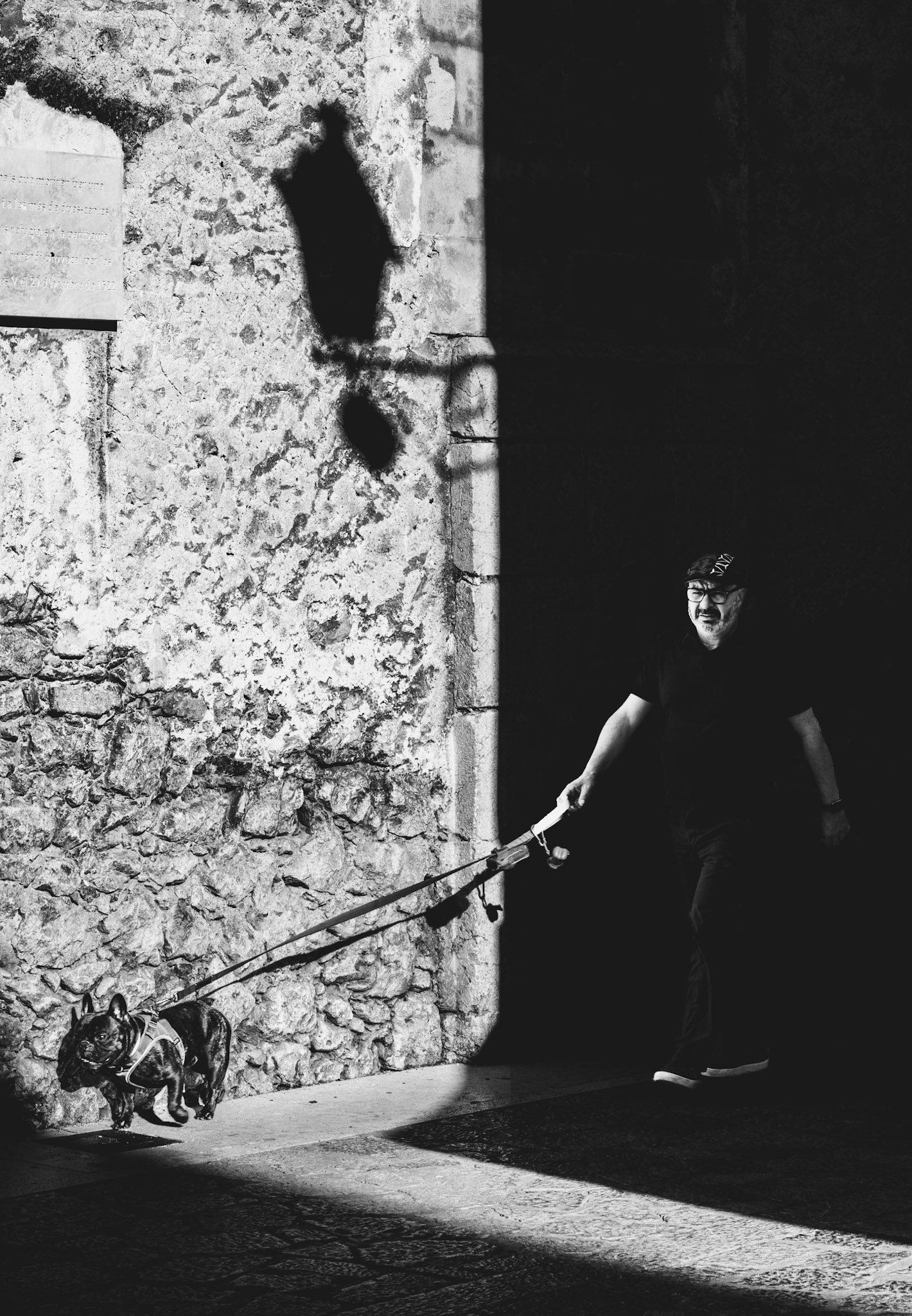 a dog pulls eagerly on its leash, its leap appears as if it's being drawn into the light. the man leading it steps half out of the deep shadow into the bright light that spills over the old stonework. the scene speaks of an unexpected discovery, almost as if a hidden moment is dragged into the light. the interplay between light and dark across the rough stones and the smooth street captures the contrast between concealed and revealed moments — the balance of light and shadow, visible and invisible, known and unknown.
