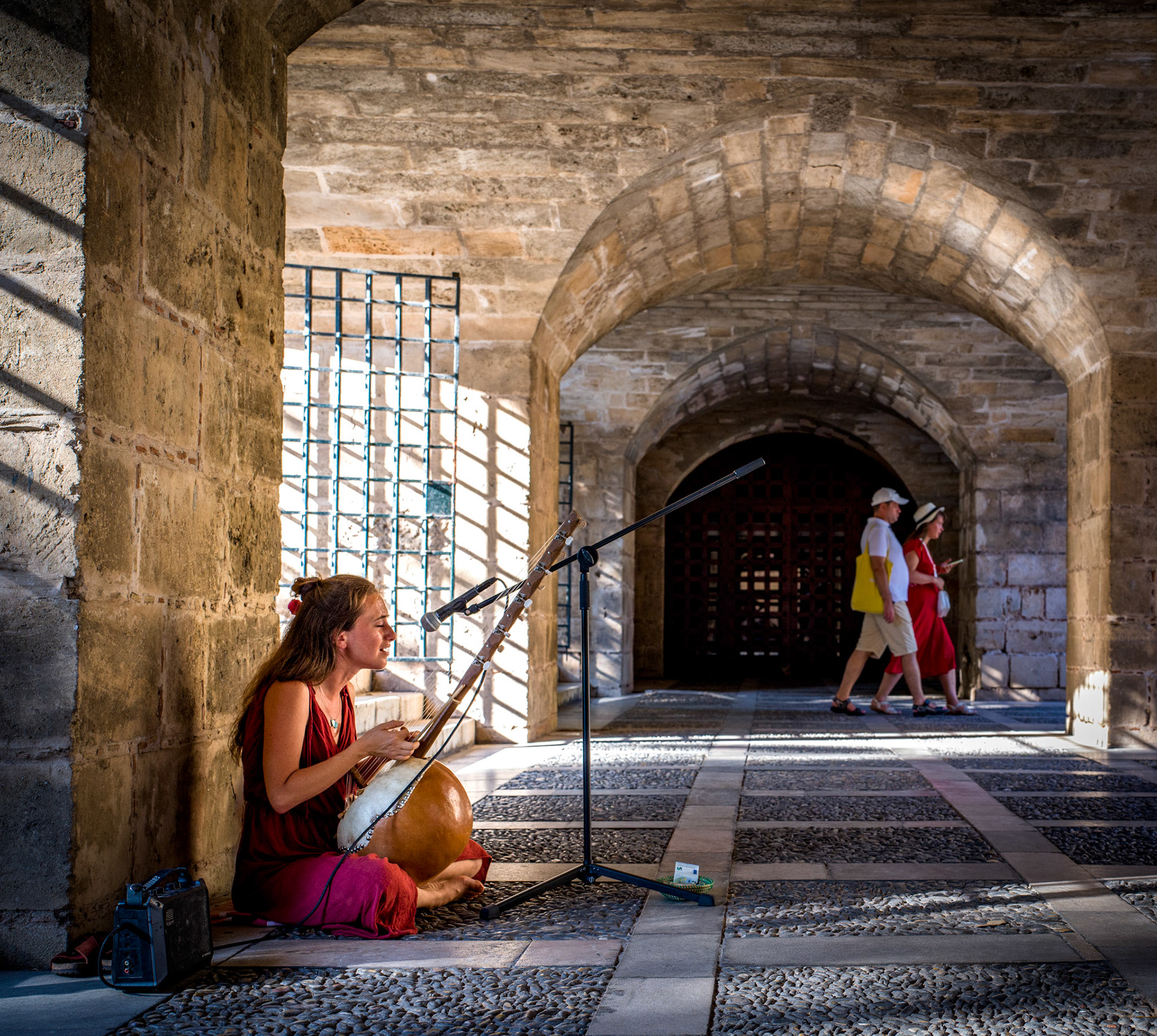 captured during a serene evening between the paseo del borne and the cathedral of palma, this image showcases a street musician playing a traditional kora, a west african stringed instrument. the warm light filtering through the stone arches adds to the tranquil atmosphere, perfectly complementing the melodic sounds of the kora.