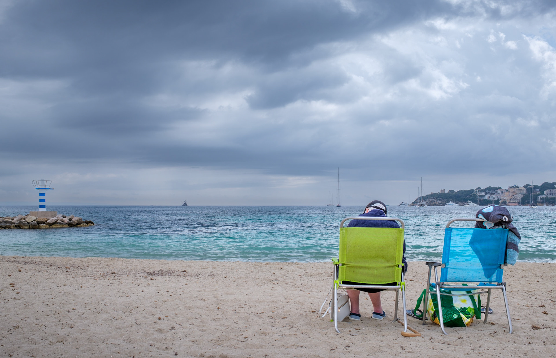 on the serene shores of palmanova, just before the season's crowds and rains descend, a tranquil moment is captured. two chairs face the open sea, one occupied, one waiting—symbols of shared solitude and anticipation. the overcast sky, a canvas of soft greys, mirrors the calm yet moody waters, setting a scene of quiet contemplation. a lighthouse stands guard at the edge, a beacon of stability in the fluid landscape. this photograph, with its blend of stillness and subtle tension, evokes the quiet before the storm, a reflective pause in the rhythmic ebb and flow of coastal life.