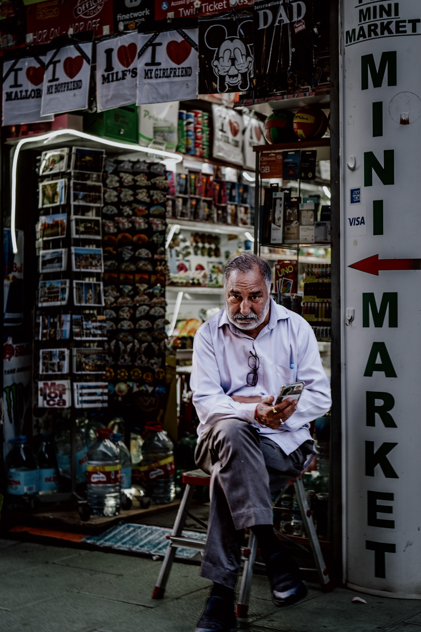 amidst the colorful chaos of postcards, souvenirs, and playful t-shirts, he sits quietly on his stool, disconnected from the hustle around him. his worn face, softened by time and habit, contrasts with the vibrant goods on display, yet there’s an unspoken connection between him and his surroundings—a man whose life rhythm has become part of this busy street corner. his phone, held loosely in one hand, seems like a window to another world, while the other hand rests gently on his knee, grounding him in the present moment. a life spent witnessing the rush of fleeting tourists, all while remaining still in the flow of the city.