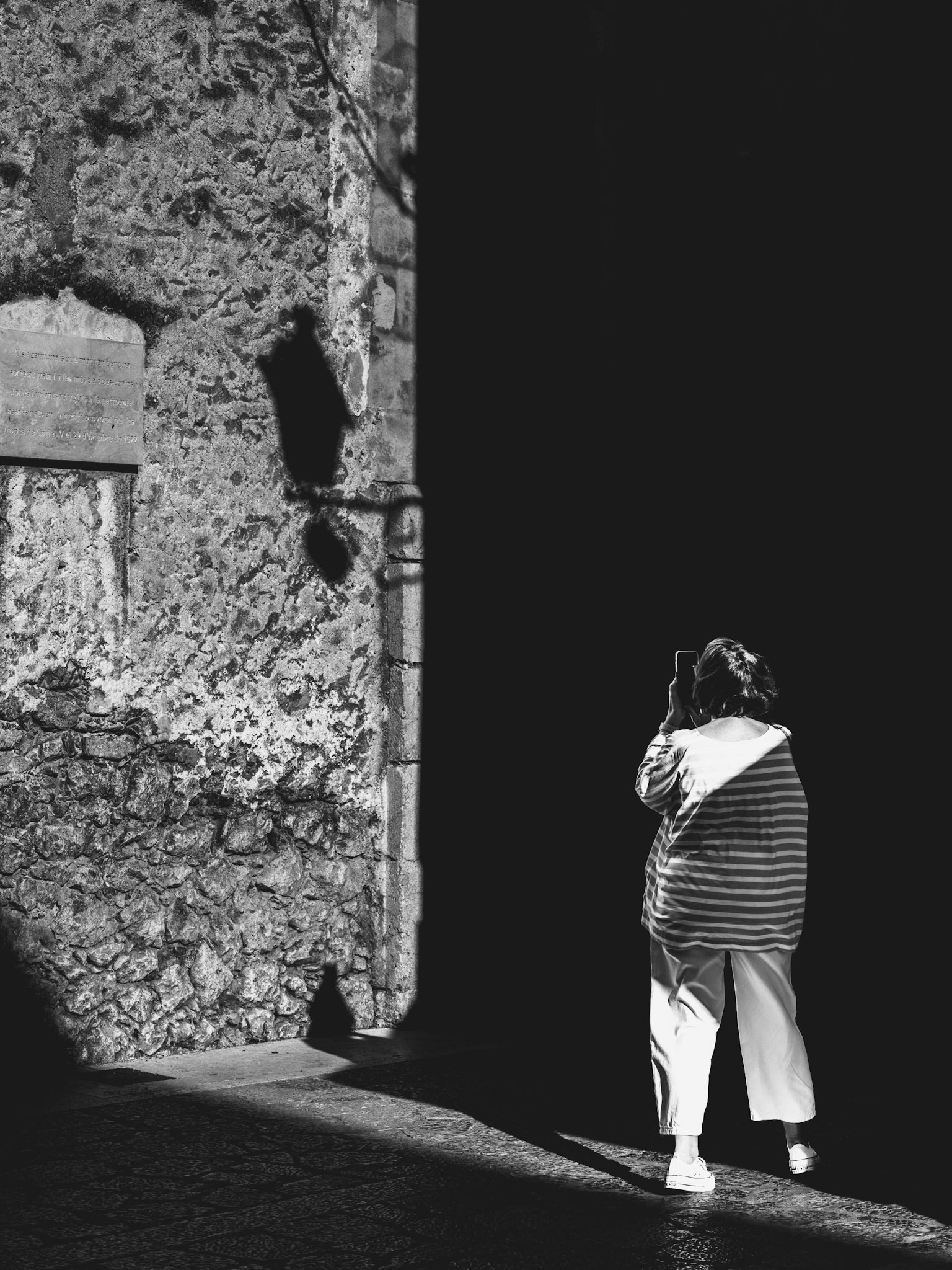 the sun slices through the old town of pollença, painting sharp edges of light and shadow. a woman stands against the stone wall, caught in the shimmering cross-light, her phone raised, her face hidden in the dark. she captures what i also see – a symbiosis of forms, a silent hunt for moments. the streetlamp casts a double shadow, a parallel narrative in space. the wall tells its story through centuries-old grooves and cracks. everything is here, held in a fragment, a picture within a picture, a photograph of photography.