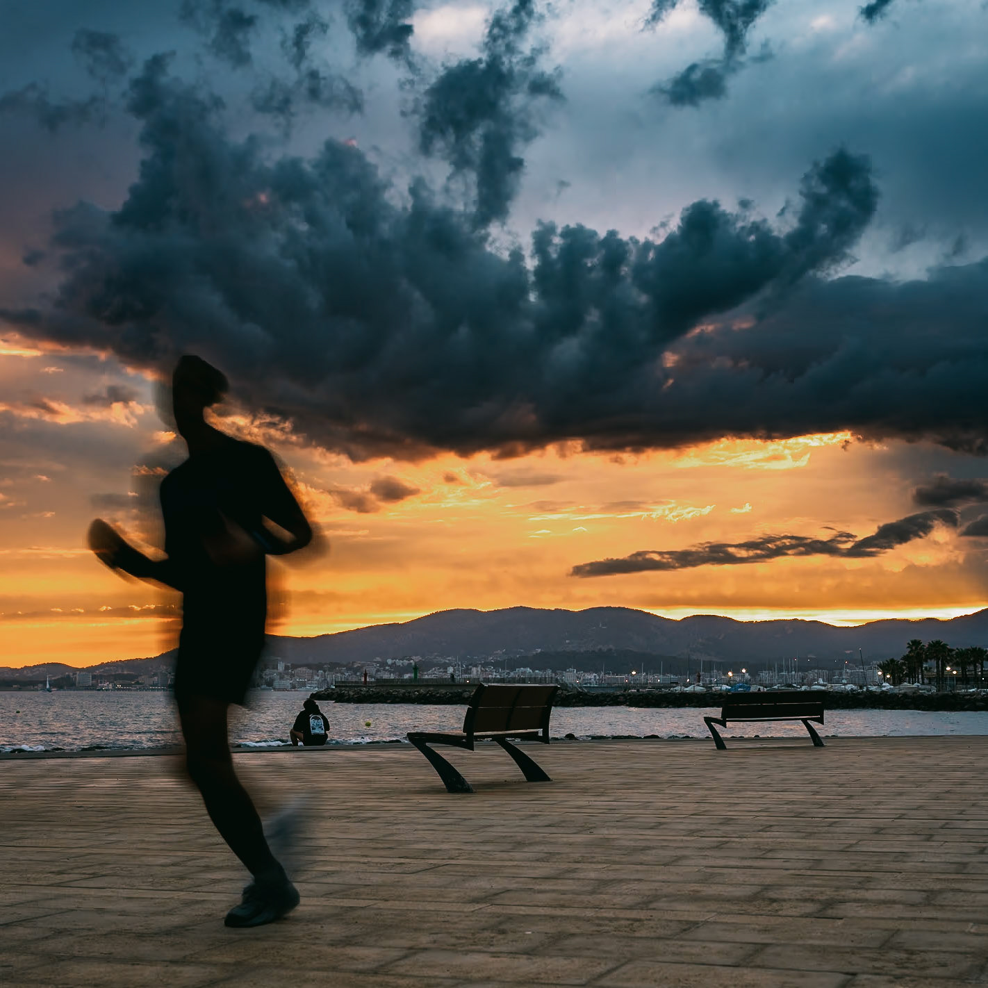 as the sun sets over portixol, casting a fiery glow across the horizon, a lone runner blurs into the scene, his form as fleeting as the day's last light. this image captures the essence of motion against a backdrop of stillness, the vibrant skies clashing with the tumultuous clouds overhead. it’s a dance of color and shadow, where the tranquility of the waterfront benches contrasts sharply with the hurried pace of the figure rushing past. the scene is a metaphor for the transient moments we often overlook, each stride a fleeting brushstroke on the canvas of time.