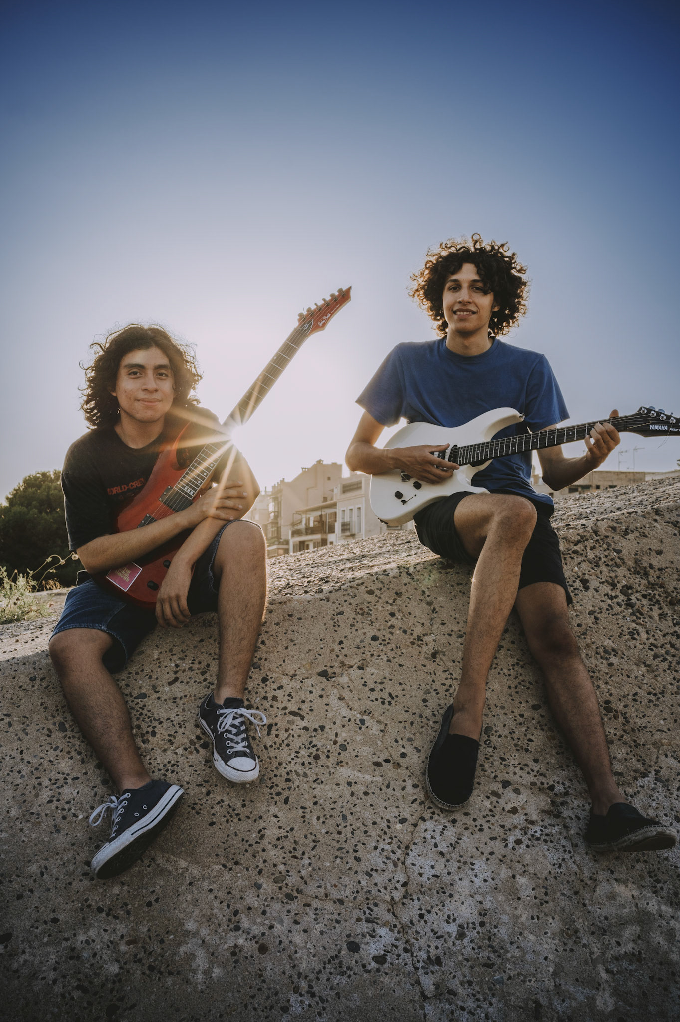 on the old stone city walls of calatrava in palma de mallorca, two young musicians sit casually, their guitars in hand, under the warm, setting sun. the sun’s rays burst through, adding a magical glow to the scene, highlighting the joy and connection between the boys and their music. the natural light captures their relaxed expressions and the effortless harmony between them and their instruments. this spontaneous moment, shot in a break from a professional session, perfectly encapsulates the spirit of youthful creativity and the simple pleasure of making music together. enhanced with a touch of brightness, contrast, and a warm color balance, this image radiates the vibrancy of the golden hour.