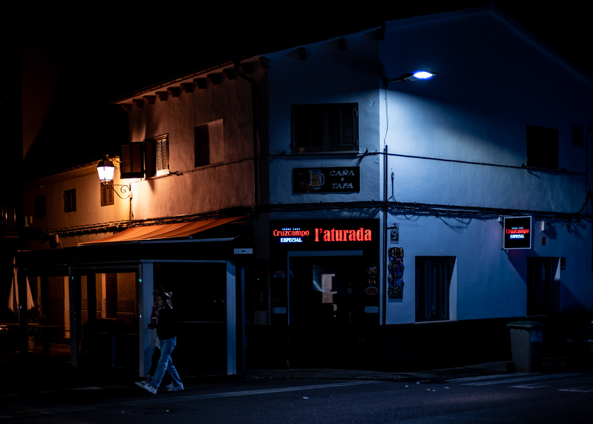 here, where the pulse of pollença's night gently throbs, two silhouettes share a fragment of time under the watchful eye of a lone street lamp. its light spills across the scene, touching the stark white walls of l'aturada, crafting an island of luminance in the sea of night. one figure stands absorbed in a distant conversation, her mobile phone a lifeline to another place, another's voice whispering secrets into the quiet of the darkened street. her companion waits, a silent sentinel, perhaps contemplating the quiet scene or lost in his own reverie. this picture captures an interlude in the nocturnal rhythm, a private bubble of connection amidst the public stage of an urban setting. it's a modern vignette, two lives intersecting briefly in the tranquility of a sleepy town, a contemporary portrait of human connection against the timeless backdrop of mallorcan architecture.
