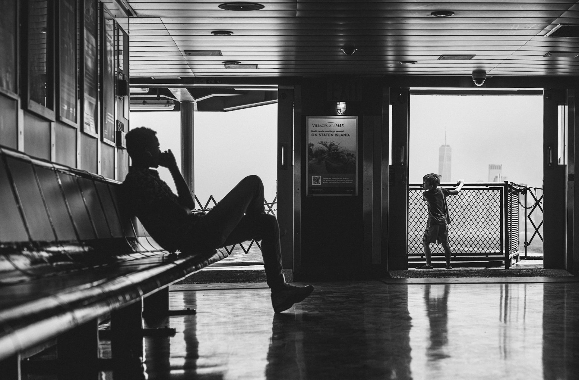 captured aboard the staten island ferry in new york city, this monochrome photo encapsulates a moment of quiet introspection amid the bustling journey. a man sits in silhouette, deep in thought, or perhaps in conversation, as a child stands by the railing, gazing out at the distant cityscape. the stark contrast between light and shadow, the reflective floor, and the framed view of the skyline create a timeless, almost cinematic feel. it’s a scene that speaks to the juxtaposition of motion and stillness, a fleeting pause in the rhythm of the city.