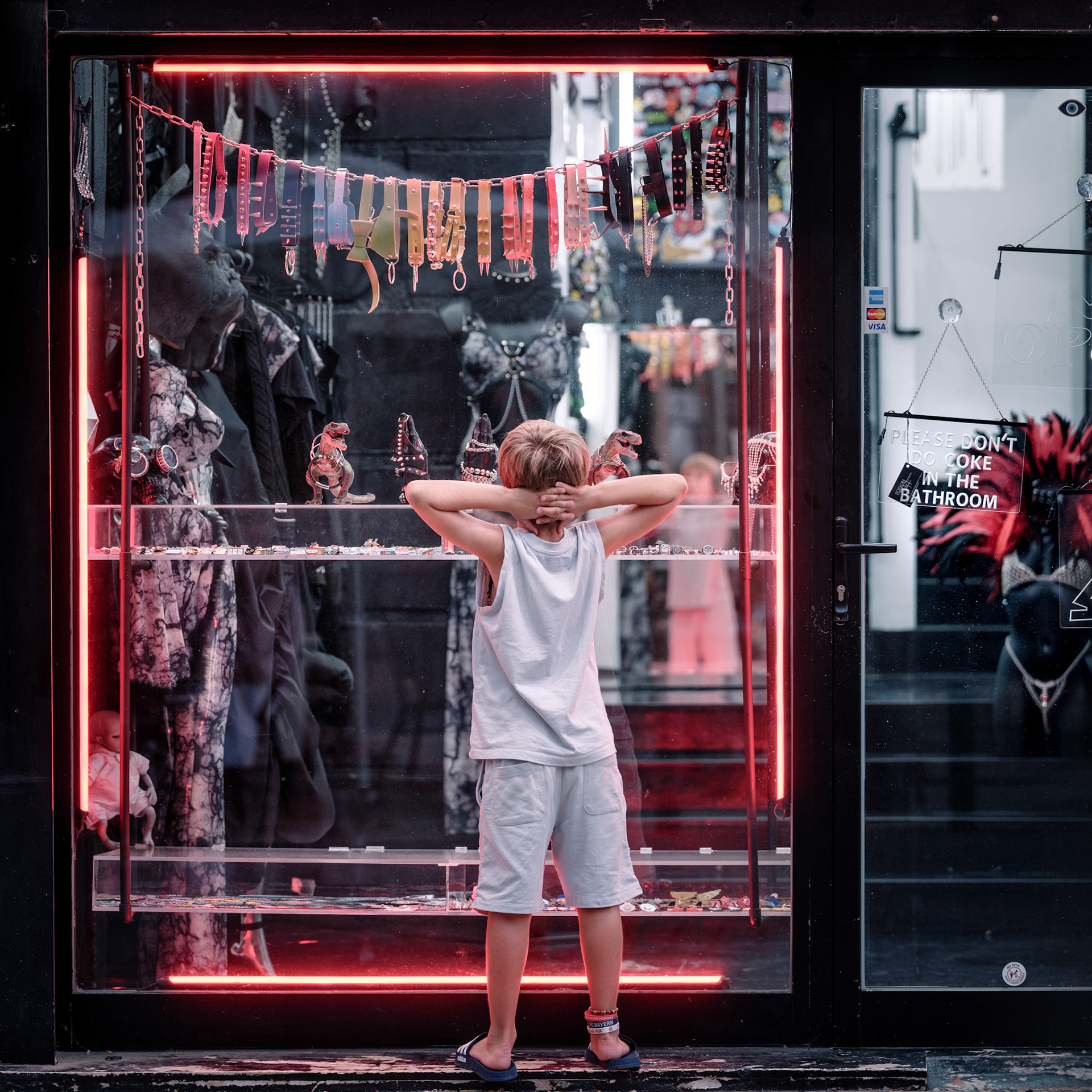 a young boy stands captivated in front of a store, his gaze fixed on the edgy trinkets and eccentric accessories inside. framed by vibrant neon lights, the shopfront offers a glimpse into a world far removed from the ordinary. inside, the shopkeeper observes, waiting for a spark of curiosity to lead the boy inside to explore a universe of bold self-expression. this is not just a store—it’s a portal to rebellion, where fashion transcends norms and becomes a statement of individuality.