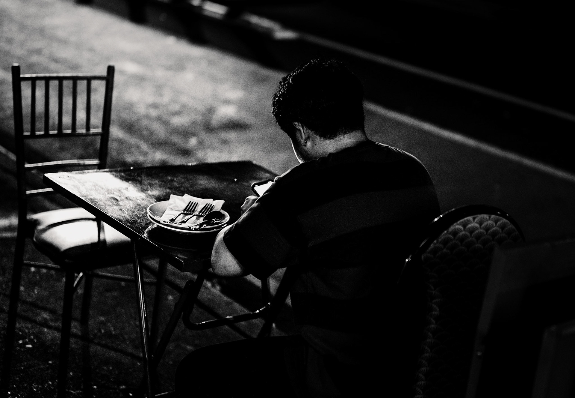 outside the restaurant soothr, on a hot summer evening in new york city, a lone figure sits at an isolated table, set apart from the rest by already folded chairs and stacked tables. whether an employee, the chef, or a solitary guest, the person is engrossed in their phone, illuminated by the soft streetlights. the image encapsulates the quiet moments of city life, where even in the hustle and bustle, pockets of solitude exist. the contrast between the darkness and the lit figure adds depth, highlighting the introspective nature of this urban scene. and by the way, the food there is indeed quite good.