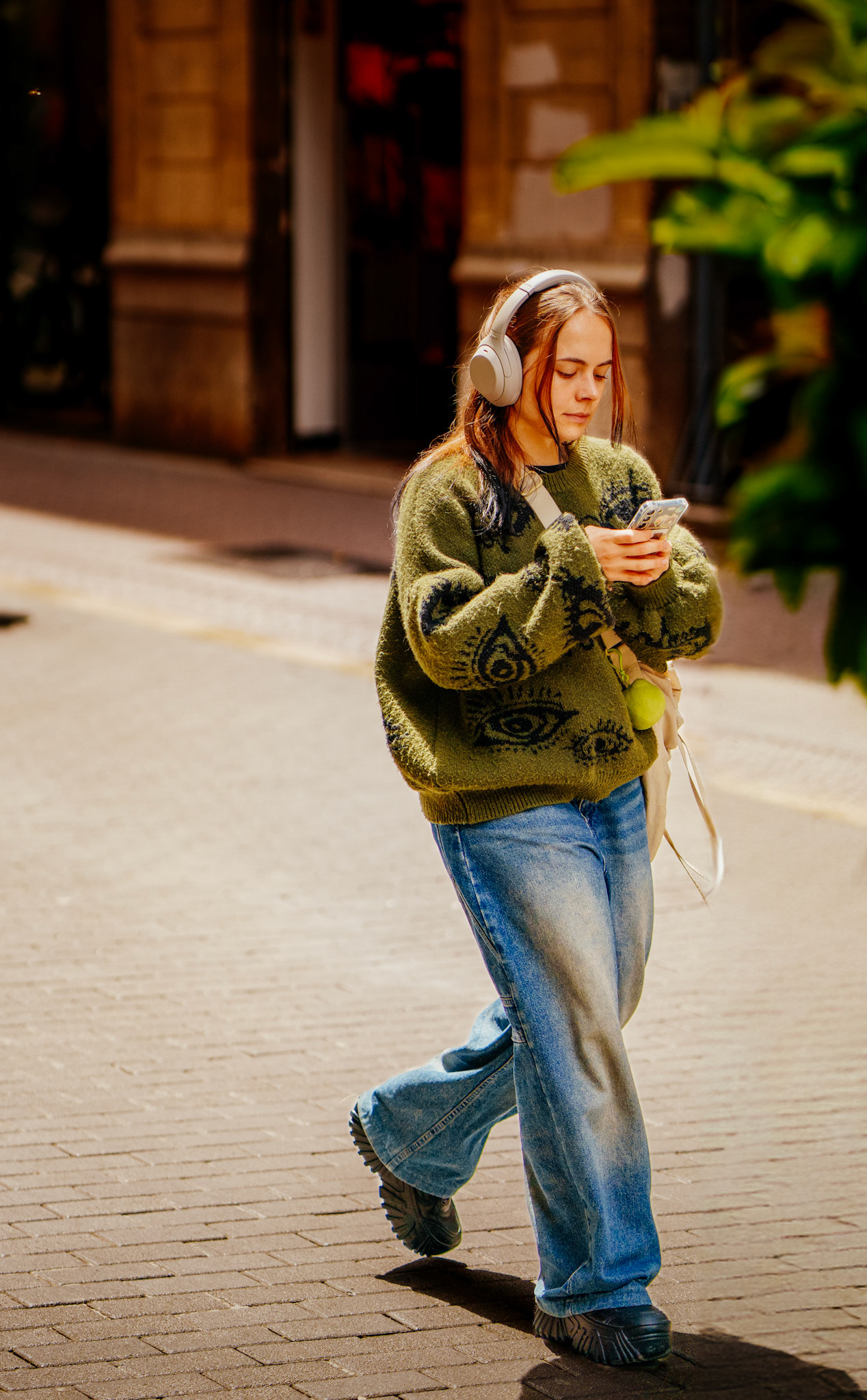 just a few rhythmic steps away from the mercat de olivar, on the path to plaça mayor in palma, she finds her own private concert. wrapped in a moss green sweater adorned with watchful eyes, she's lost in a melody that dances just for her. the soft glow of daylight caresses her contemplative face, while her hands, cradling the source of her tune, seem to be the conductors of her inward symphony. she's a soloist moving through the urban tempo, her wide denim pants catching the beat of her stride. it's a silent recital where every step is a note, every glance a lyric, in the symphony of the streets.