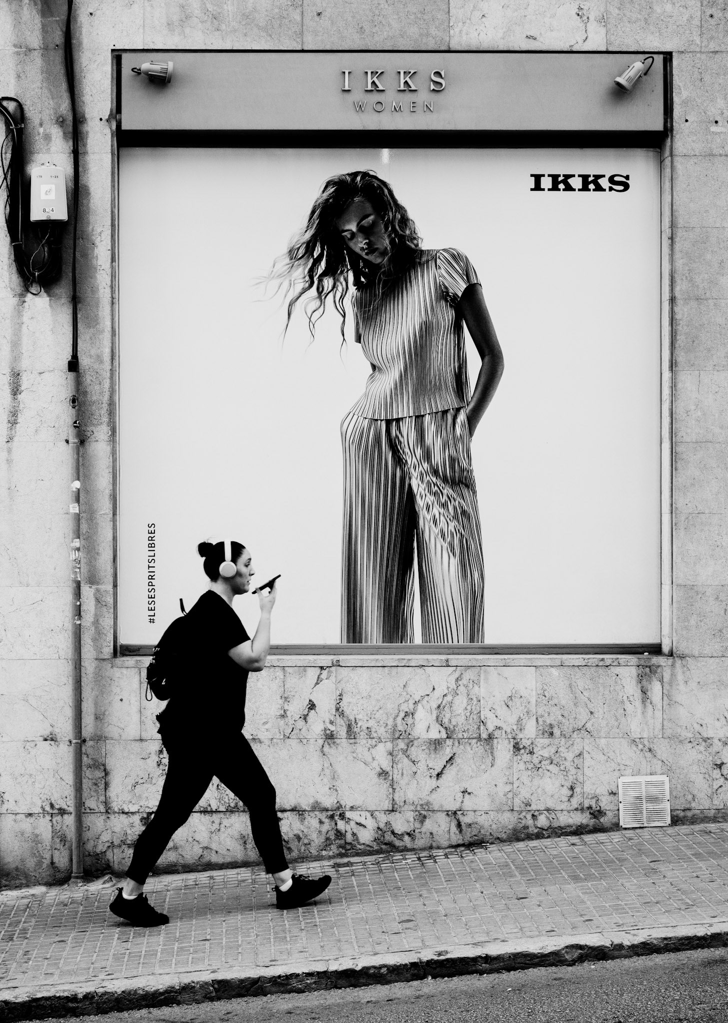 captured on a lively street in palma de mallorca, this photograph juxtaposes the movement of modern life with the timeless, frozen elegance of fashion. the woman on the sidewalk, immersed in her own world with headphones on, strides confidently past the larger-than-life poster. the ad's figure seems to lean down, almost watching or interacting with the passerby, creating a dynamic tension between the real and the imagined. this snapshot highlights the blend of the everyday with the aspirational, a dance between reality and the idealized world we are constantly presented with.