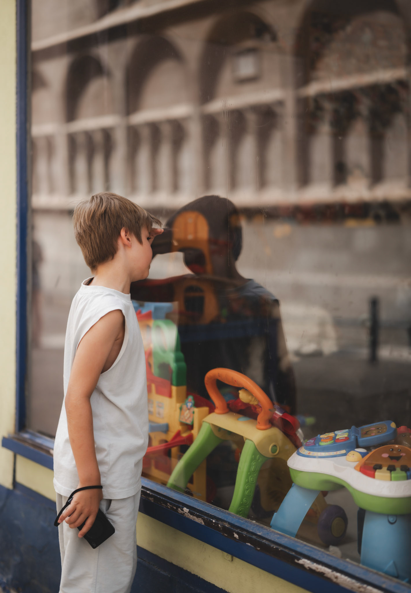 on a casual photowalk through the vibrant streets of palma de mallorca, a young boy stands captivated by a window display of colorful toys. his reflection merges with the scene behind the glass, creating a beautiful juxtaposition of childhood wonder and budding creativity. holding his own camera, he balances between the innocence of an 8-year-old and the promise of a young photographer, his eyes filled with dreams and stories waiting to be captured.