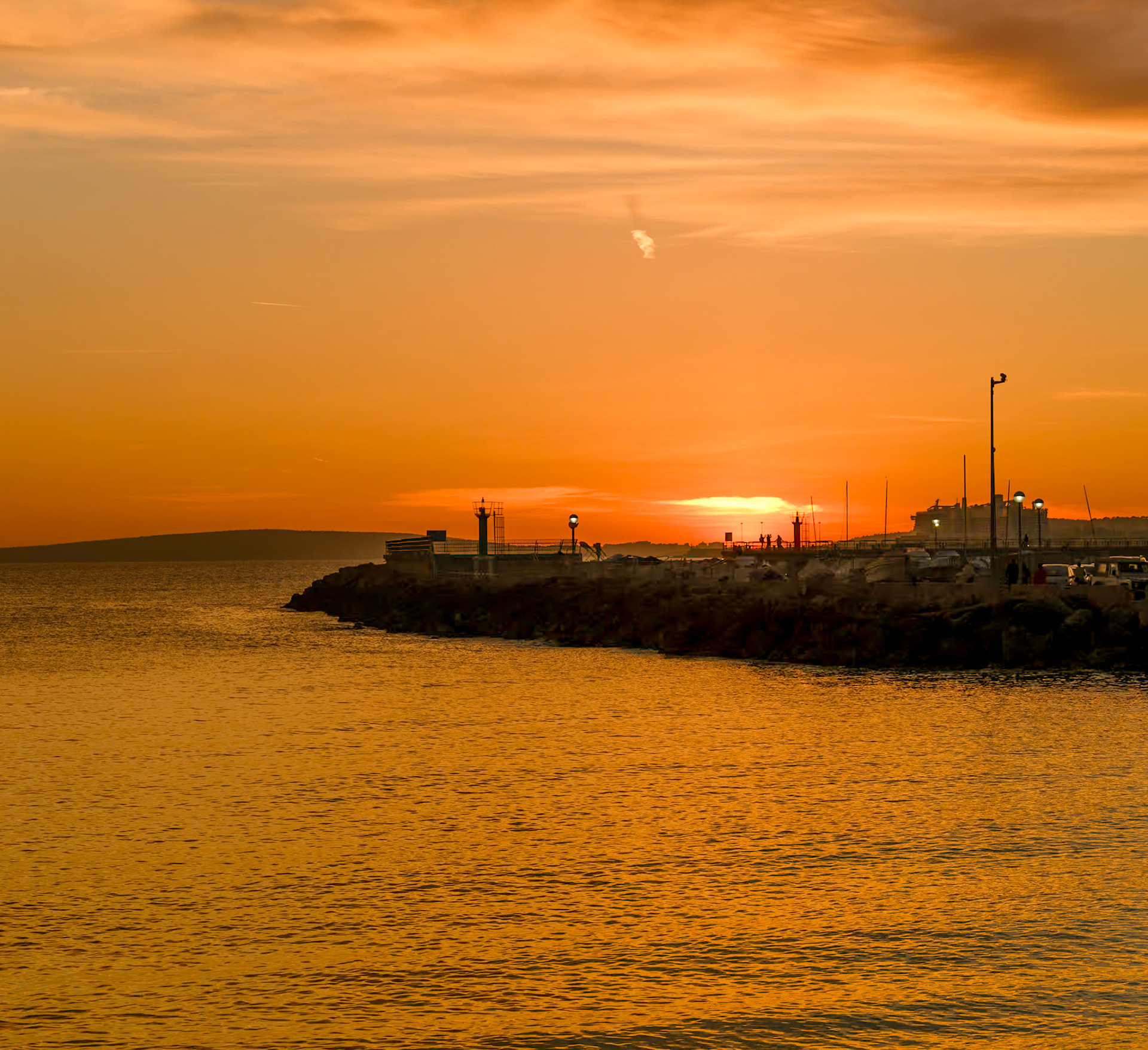 at portixol, a district of palma de mallorca that rests by the sea, the setting sun casts a golden farewell to the day. Silhouettes of people and structures are sketched against the fiery horizon, while the calm waters reflect the fading light. It's a tranquil scene where the ocean meets the sky in a warm embrace, a daily spectacle observed by locals and visitors alike, each sunset unique to the beholder.