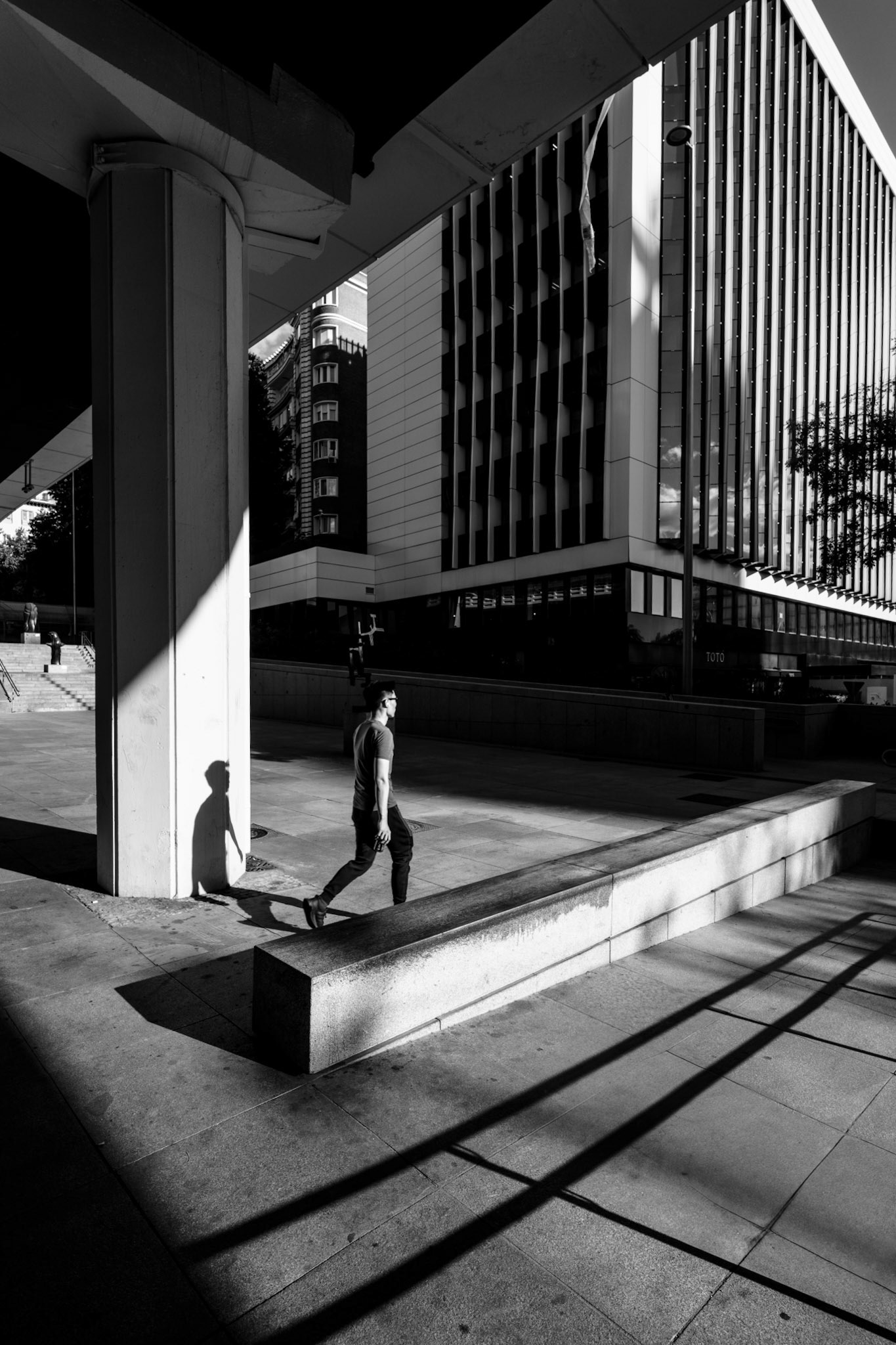 captured at the urban intersection of museo de escultura al aire libre in madrid under the causeway Eduardo Dato, where modern architecture meets the flowing lines of sunlight. a solitary man moves through the rigid structures of the city, yet his shadow stretches freely across the ground. the fractured shadow lines and sharp contrasts highlight the scene’s symmetry and dynamism, while the interplay of light and darkness reflects the duality of freedom and confinement in an urban environment.