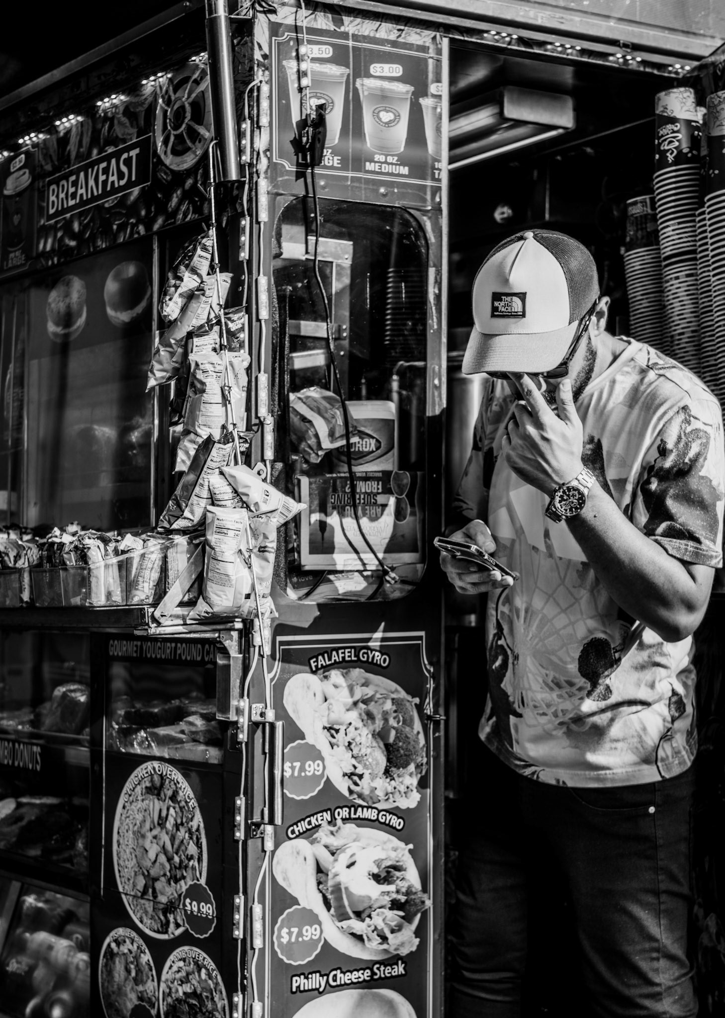 captured at the bustling corner of broadway, this candid shot reveals a street vendor taking a brief pause from serving customers at his vibrant food cart. bathed in the glaring sunlight, he squints at his phone, perhaps checking messages or scrolling through social media. the food cart, adorned with colorful signs advertising falafel gyros and philly cheese steaks, adds a rich backdrop of urban life. this photo encapsulates a fleeting, yet intimate moment of stillness amidst the constant rush of new york city, highlighting the daily grind and the small moments of rest that punctuate it.