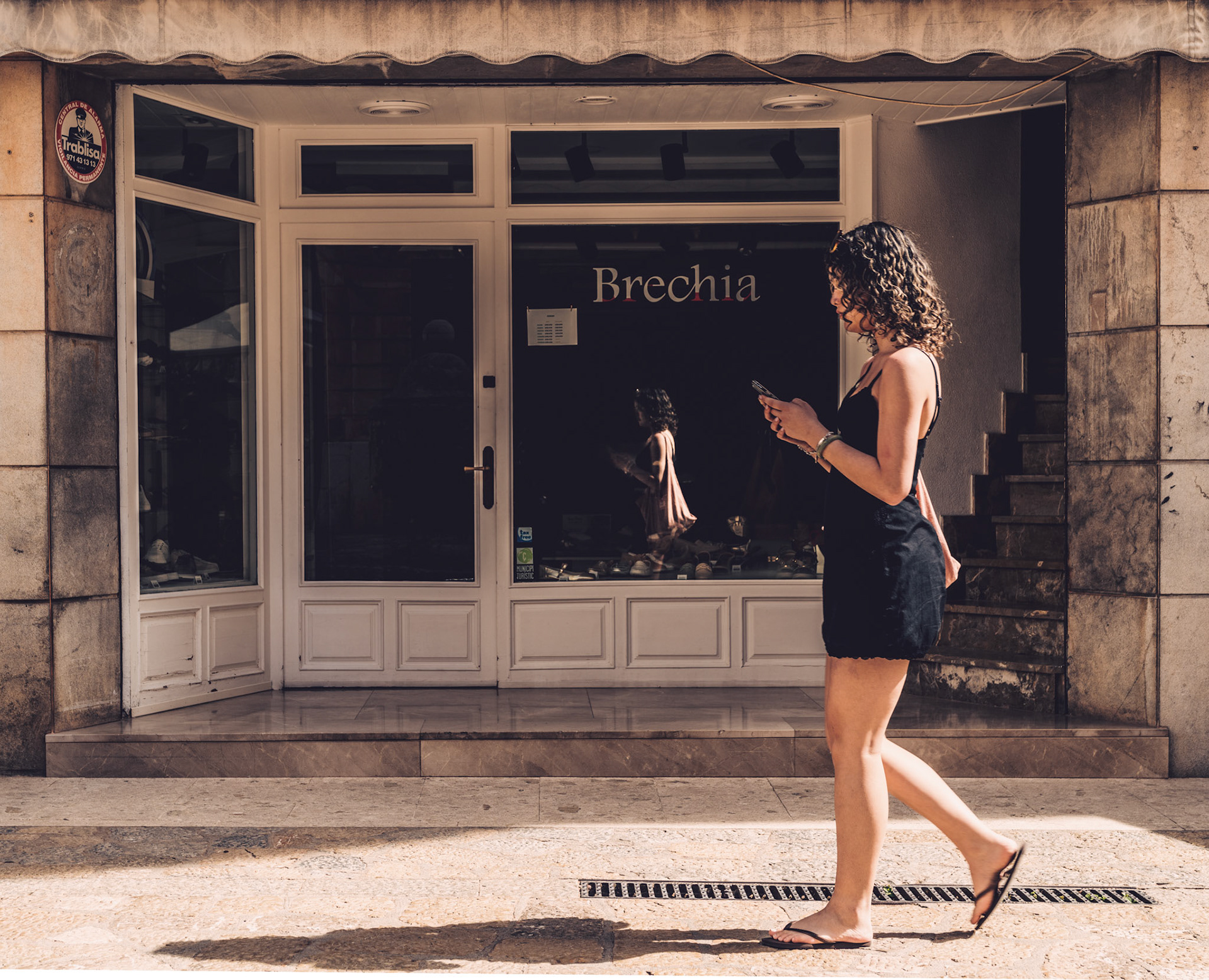 strolling through the old town of pollença as the late afternoon sun washes the cobblestones in a warm glow, here's a moment where two worlds gently collide. the young woman, engrossed in her digital world, walks by a storefront named 'brechia', unaware of her reflection that perfectly mirrors her in the shop’s glass— a silent twin in the physical realm. it's a dance between the tangible and the transient, a snapshot where technology intersects with tradition in the heart of mallorca’s northeastern gem.