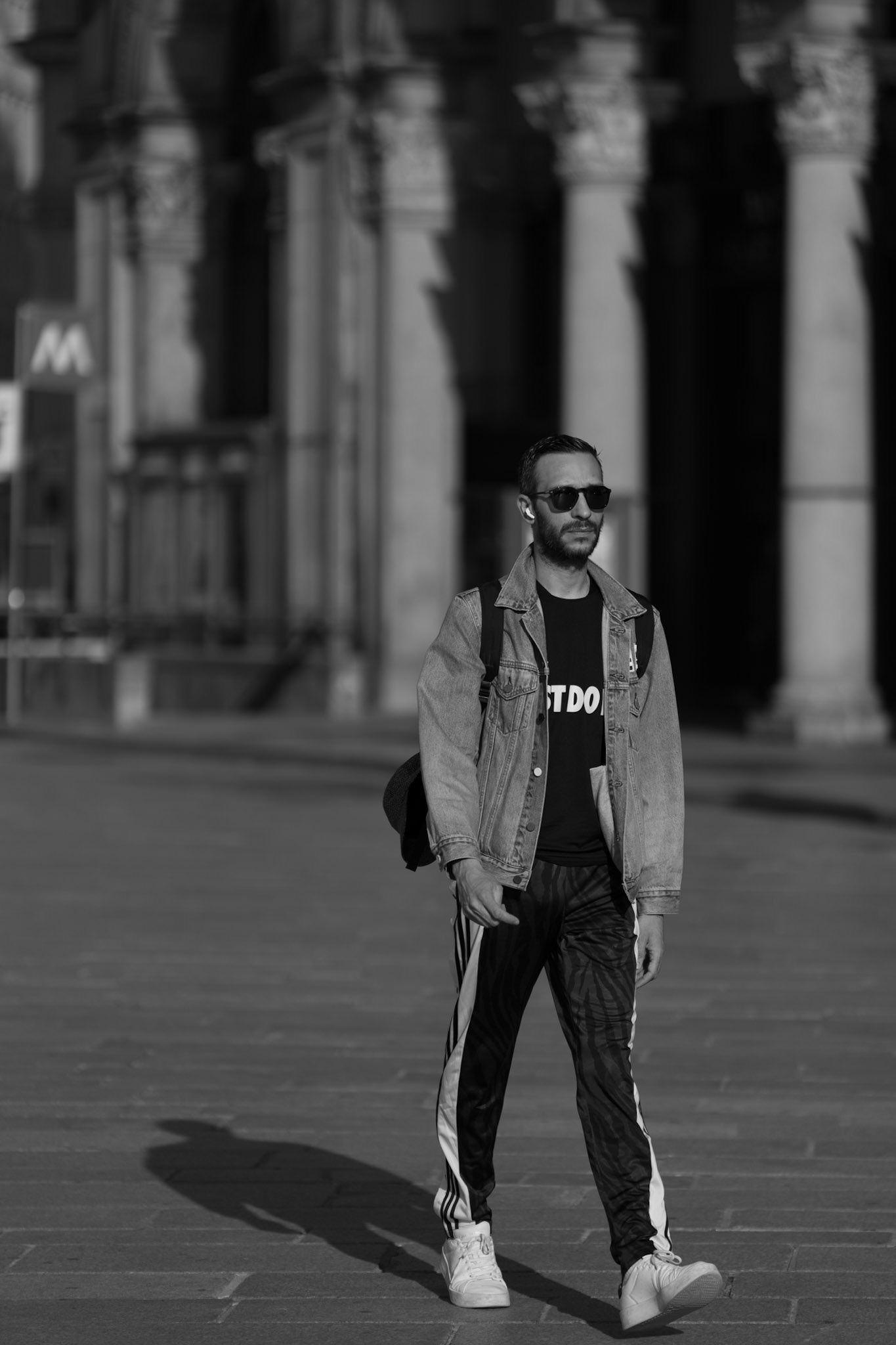 Captured in the iconic streets of Milan, this black and white photograph depicts a man in mid-stride, exuding confidence and a cool, contemporary style. His outfit—a classic denim jacket paired with athletic stripes and casual sneakers—perfectly mirrors Milan's reputation as a fashion hub. The play of shadows and architecture in the background adds depth and a sense of movement to the image, emphasizing the city's dynamic and historical essence. His sunglasses and composed demeanor give him an air of mystery, making this more than just a momentary snapshot; it's a portrayal of a lifestyle, blending casual comfort with urban sophistication in Milan's ever-moving landscape.