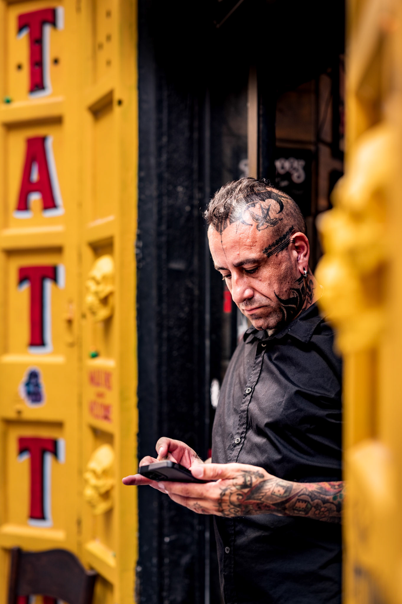 this candid street portrait captures a tattoo artist taking a moment's pause in the doorway of his studio in la lonja, palma de mallorca. the vibrant yellow of the door contrasts with the artist's black attire and intricate tattoos, creating a visually striking composition. absorbed in his phone, the artist's demeanor conveys a blend of intensity and calm. after this candid shot, i approached him for a more formal portrait, adding a personal connection to this spontaneous encounter. the image highlights the intersection of urban life and personal artistry, encapsulating a moment of quiet reflection amidst the vibrant city backdrop.