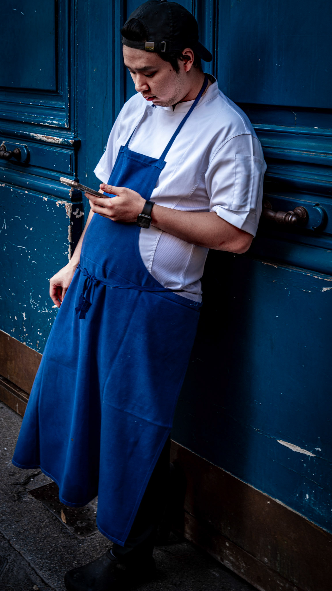 in the quiet moments away from the kitchen chaos, a chef in his crisp, blue apron takes a brief pause on the streets of paris, just near place blanche. his posture is relaxed, the phone in hand perhaps bringing news or simply offering a moment of disconnect. the worn texture of the blue door behind him contrasts with the stillness in his expression, a subtle reflection of the bustling world just outside the frame. the atmosphere captures a blend of resilience and reflection, a fleeting pause in a long day.