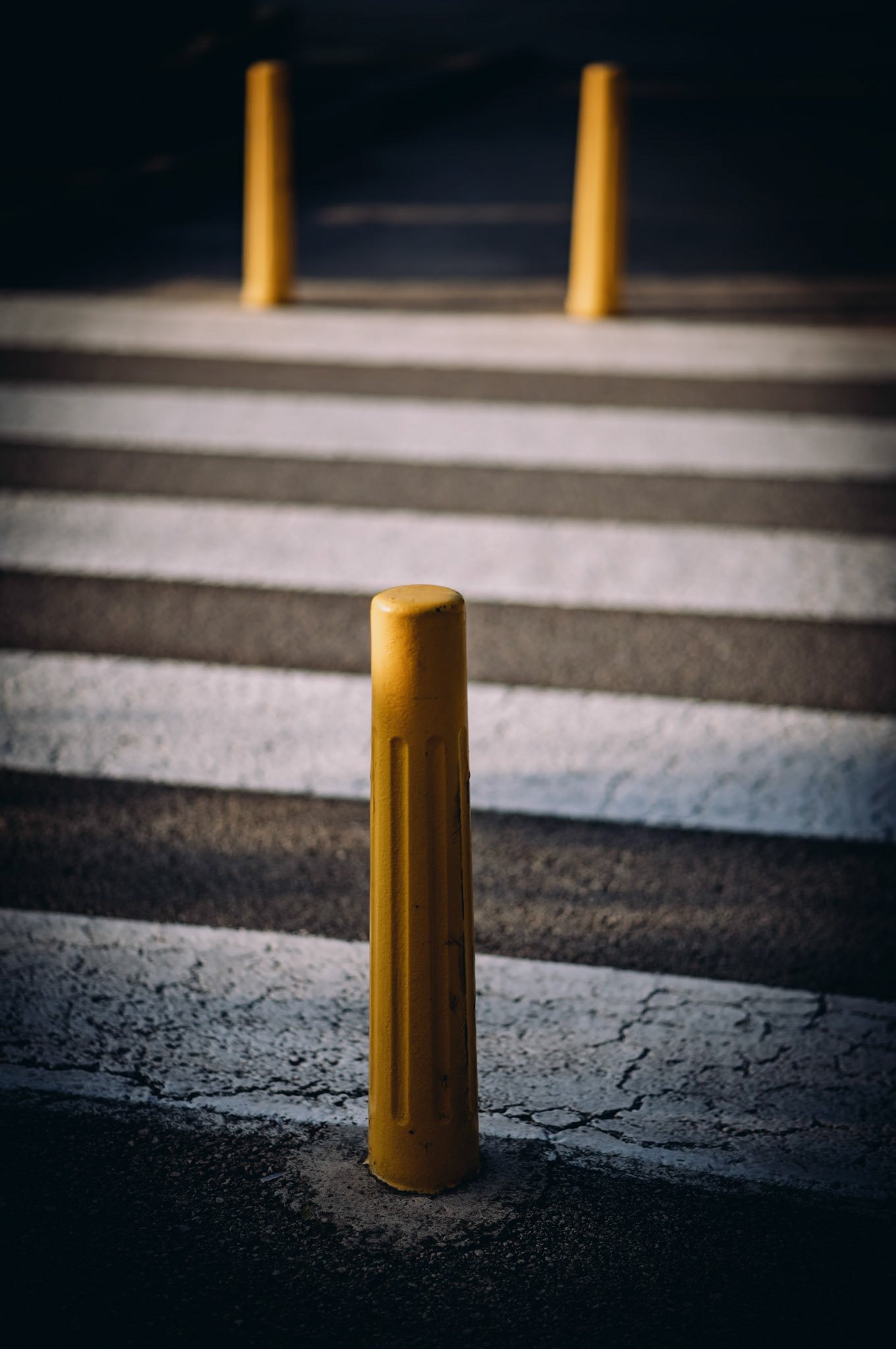 shot in alcudia, a beautiful historic village in the north of mallorca. a solitary yellow bollard stands on a crosswalk, sharply contrasting with the grey pavement. shadows and light create a geometric pattern, emphasizing the bollard's presence. the background blurs into soft focus, giving the scene a sense of urban stillness.