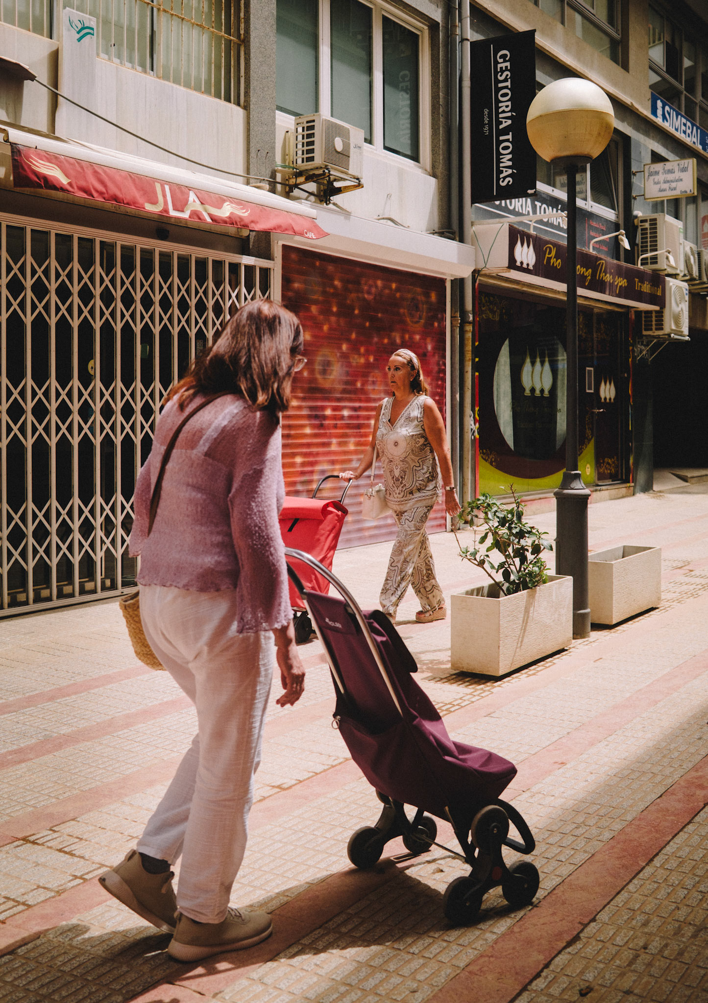 captured in a quaint, old shopping area near the mercat de l'olivar in palma de mallorca, this image humorously portrays two women seemingly "meeting" without acknowledging each other. their contrasting outfits and matching red carts add an element of symmetry and irony to the scene. amidst the bustling backdrop of closed shops and the midday sun casting sharp shadows, this moment freezes the silent choreography of urban life, where paths cross yet remain parallel.