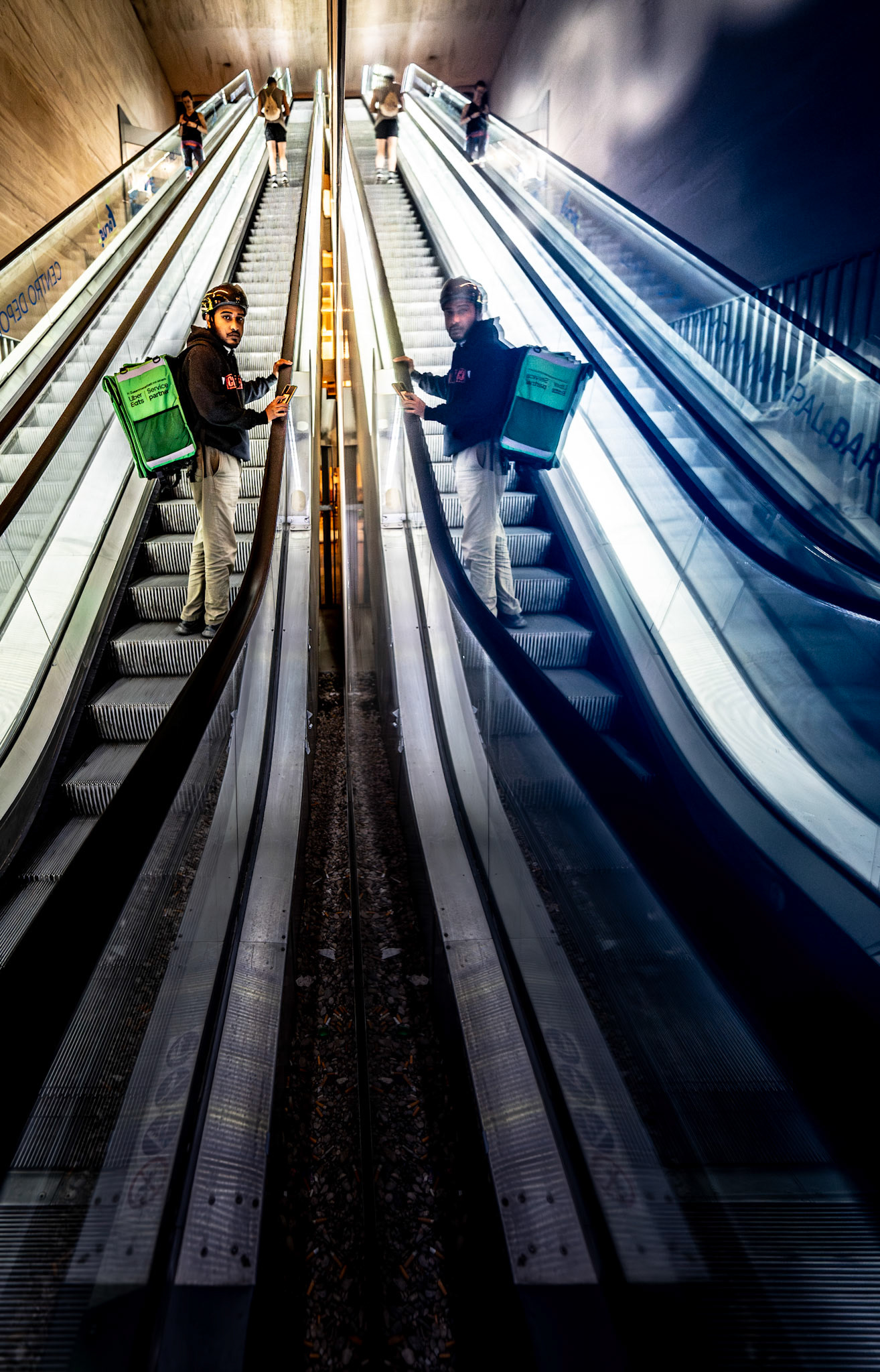 captured at madrid’s mercado barceló, where the mirrored surfaces of the escalators reflect the rhythm of urban life. the delivery rider, pausing with his phone, seems caught between movement and stillness, a brief moment of pause amid the constant flow. the cool tones and stark reflections create a cityscape that feels both intimate and infinite, where every step is mirrored by another.