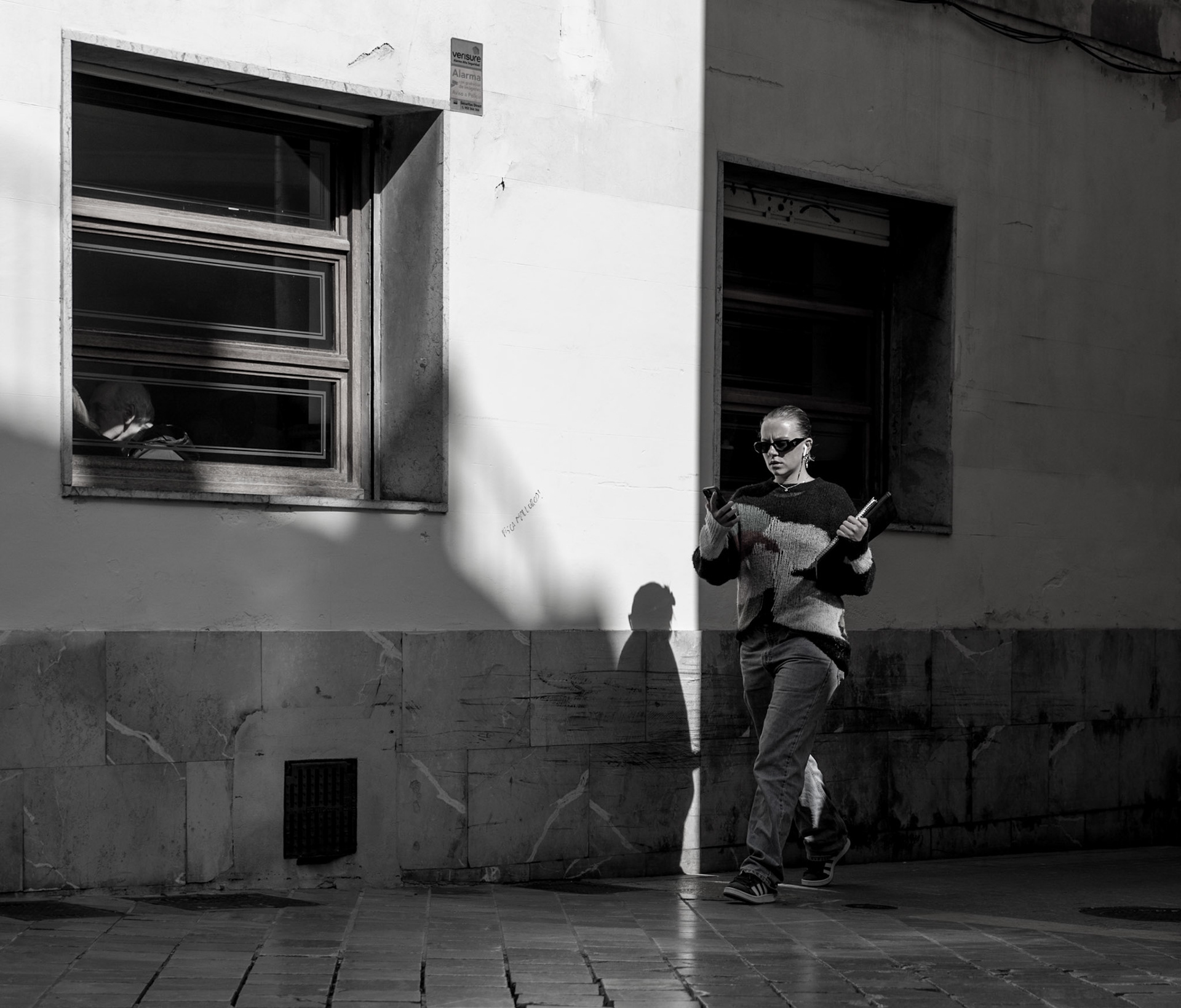 in the late afternoon light that sweeps through palma's old town, a woman pauses in stride, phone in hand, perhaps deciphering a message or seeking direction. the contrast is stark — her form illuminated in the sun's golden assertion, the shadow a quiet echo on the ancient walls. there's a rhythm here, an urban ballet, where each pedestrian is both dancer and choreographer, navigating the city's storied corridors. as the window casts its gaze and the world outside hustles by, she finds a moment of still, her attention captured by a glowing screen, a modern compass amidst the echoes of history.