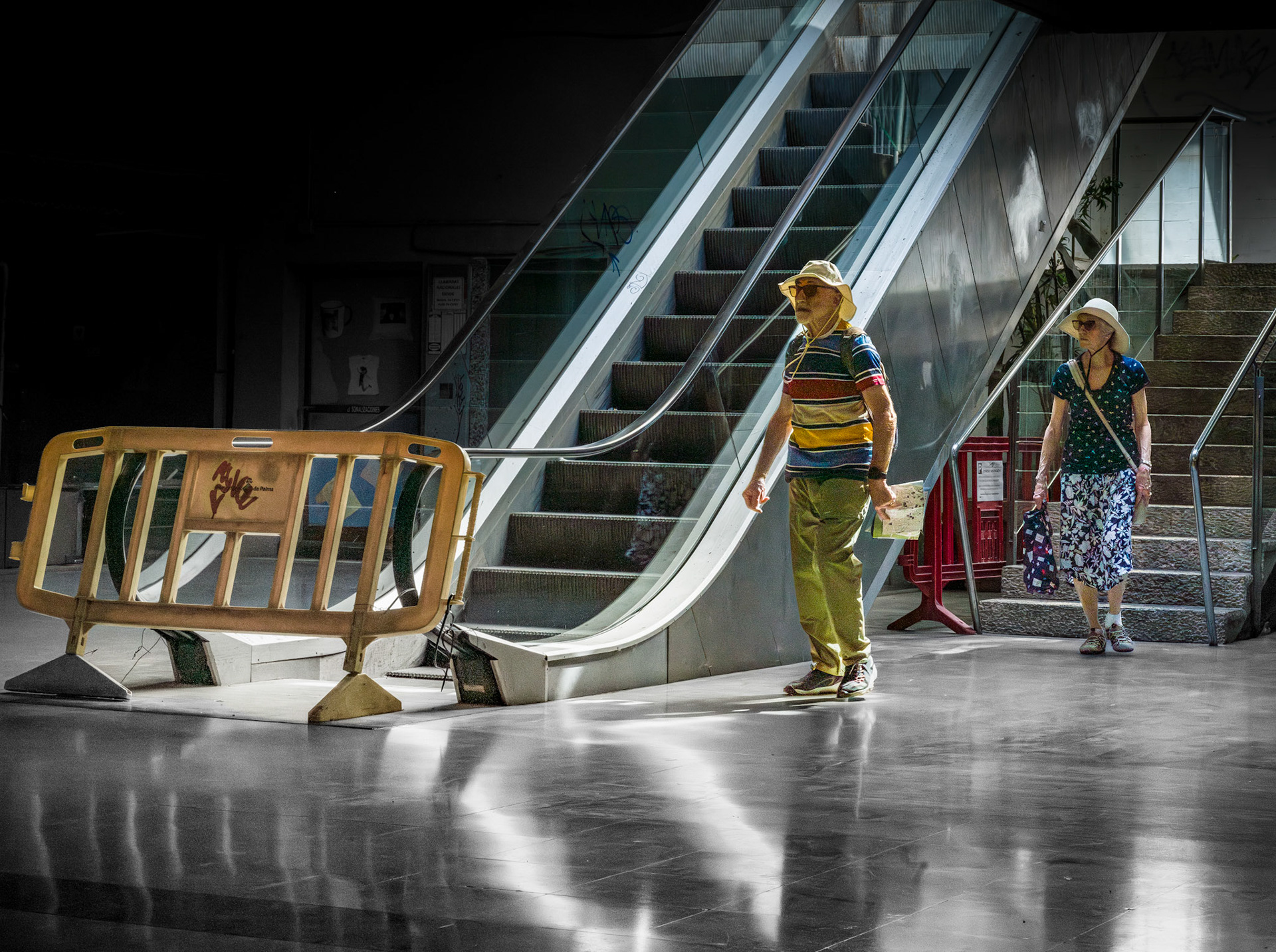 beneath the bustling plaça mayor in palma, a quiet, almost eerie scene unfolds. once home to a shopping center, this now deserted space feels like a forgotten corner of the city. tourists, like this couple, only wander here on their way to the underground parking, often unsure of their surroundings in this maze-like, dimly lit passage. the mix of the sterile, urban environment and the isolation of the space creates an intriguing tension — a hidden world beneath the vibrant streets above. the faded escalators and the misplaced construction barrier add to the feeling of abandonment, while the soft light reflecting off the floor gives the moment an otherworldly calm.