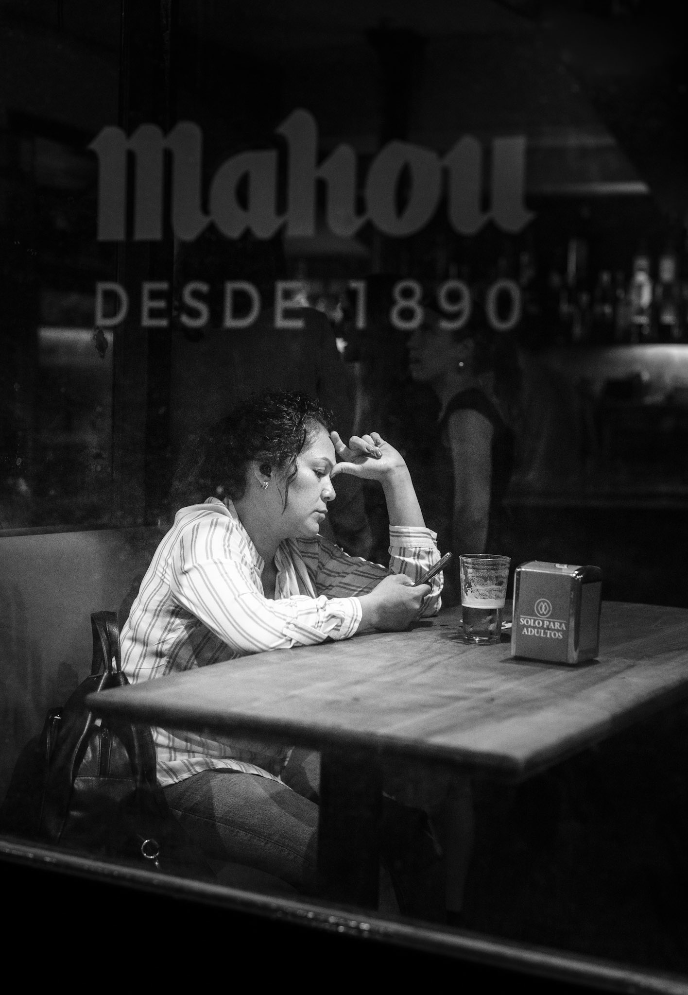 through the glass of a madrid bar, under the old script of “mahou desde 1890,” she sits alone, her fingers lightly touching the screen of her phone. her face, framed by curls, shows signs of quiet contemplation. a half-finished beer rests before her, untouched in the warmth of the dimly lit space. there is a solitude in her posture, a pause amid the sounds of conversation and clinking glasses, an urban moment suspended between the past and the present, where stories are untold, but emotions are felt deeply.