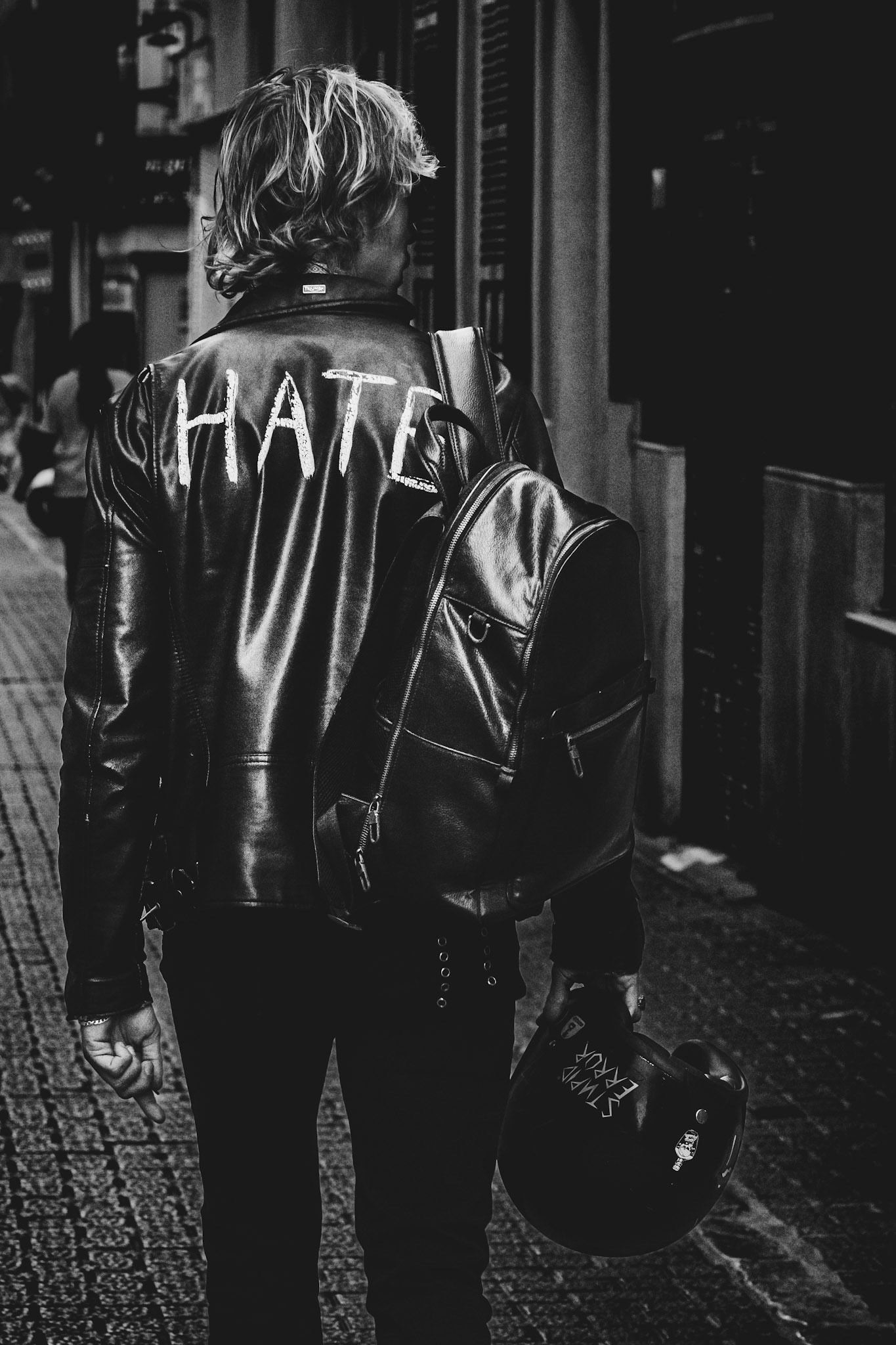 captured on a quiet street in palma, this striking image tells a story of rebellion and isolation. the "hate" boldly written across the back of the leather jacket commands attention, while the motorcycle helmet casually held in hand adds a layer of complexity, hinting at a journey or a moment of pause. walking down the narrow carrer de can vatlori, the figure is framed by the urban landscape, yet disconnected from it — a lone traveler in a world that seems distant, cold, and indifferent. the black-and-white treatment enhances the raw texture of the leather and the gritty street, creating a mood that is both intense and introspective. it’s a reflection of urban life, where emotions can be just as stark and unfiltered as the streets themselves.
