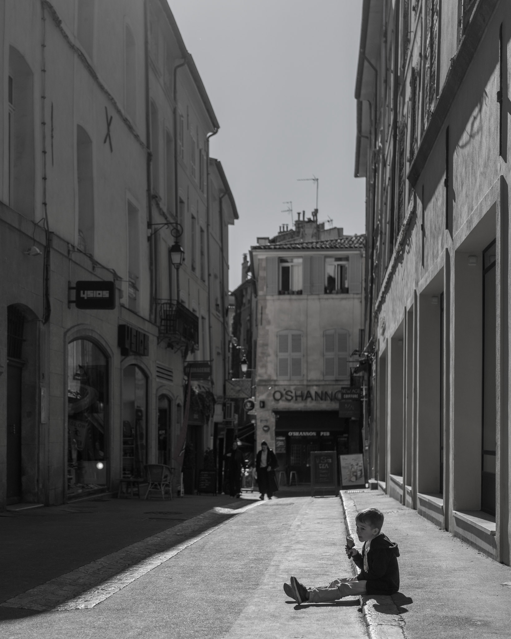 this monochromatic scene whispers a tale of simple joys amidst the grand narrative of the city. a young boy, seated on the curb of aix-en-provence's storied streets, finds a universe in the spirals of his ice cream. around him, the city breathes in the routine of daily life, unaware of the profound moment unfolding in its midst. as light carves through the alleyways, it spotlights the child, his delight casting a shadow long enough to blend with the cobblestones, telling of timeless pleasures in the transient afternoon.