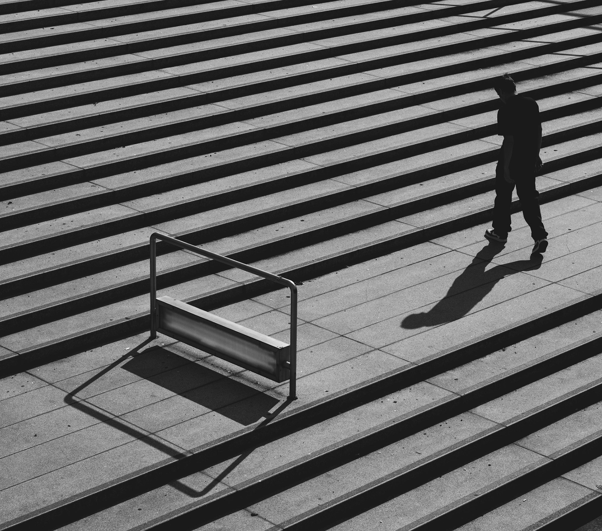 a lone figure descends the stairs of the Riem Arcaden in Munich, moving between light and shadow. each step marked by a pattern of lines and shapes, the stark contrast highlights the interplay between architecture and human presence. the solitude of the figure, casting a shadow that stretches behind like a silent companion, evokes a quiet narrative of reflection and introspection. the scene, caught in black and white, feels both timeless and immediate, drawing us into a moment where geometry meets the human experience.