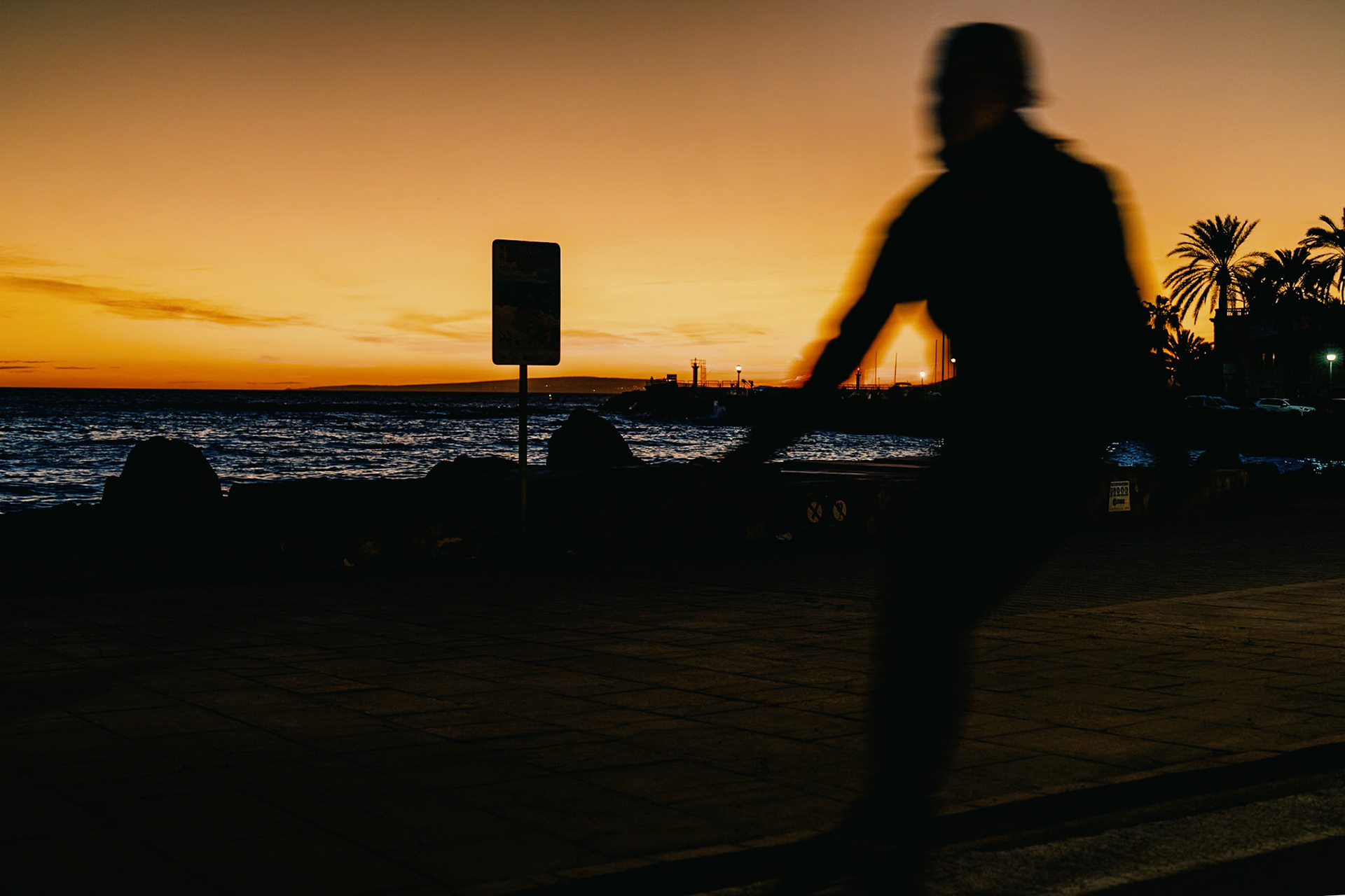 i captured this serene sunset at portixol, a vibrant district of palma de mallorca. Vivid orange and yellow hues streaking across the sky. The sun dips near the horizon, silhouetting palm trees and a distant lighthouse. A blurred figure in motion adds life to this tranquil seascape, where water meets a fiery sky.