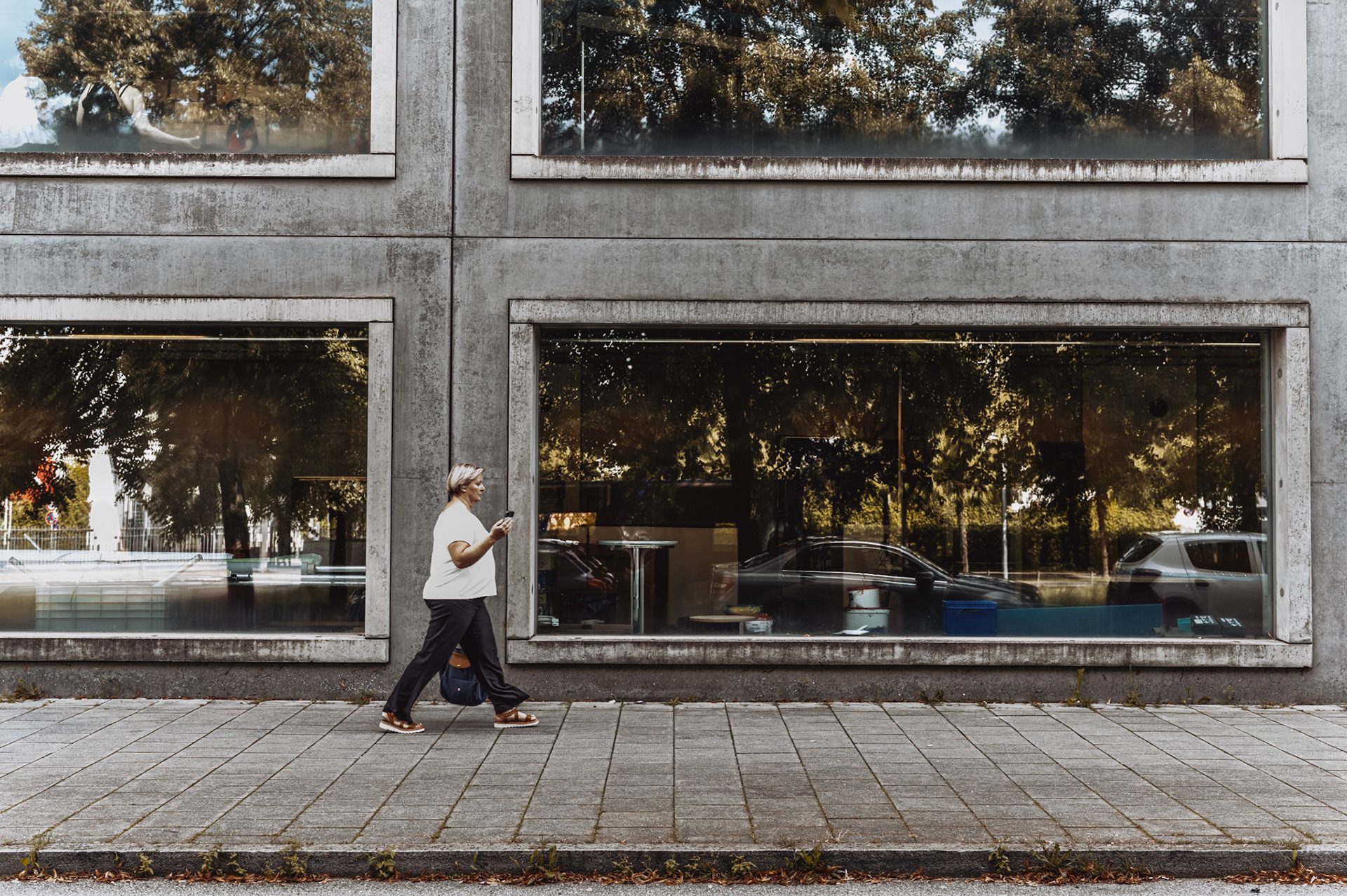 this photo captures a serene moment in münchen’s messestadt riem, where urban lines meet a hint of nature. the woman walking along the sidewalk, absorbed in her phone, contrasts beautifully with the reflected trees in the building’s large glass windows. these reflections create a “framed forest” within the concrete grid, blending the natural and the urban in a unique, balanced composition. it’s a quiet scene that finds poetry in everyday life, showing how city structures can hold unexpected glimpses of nature.