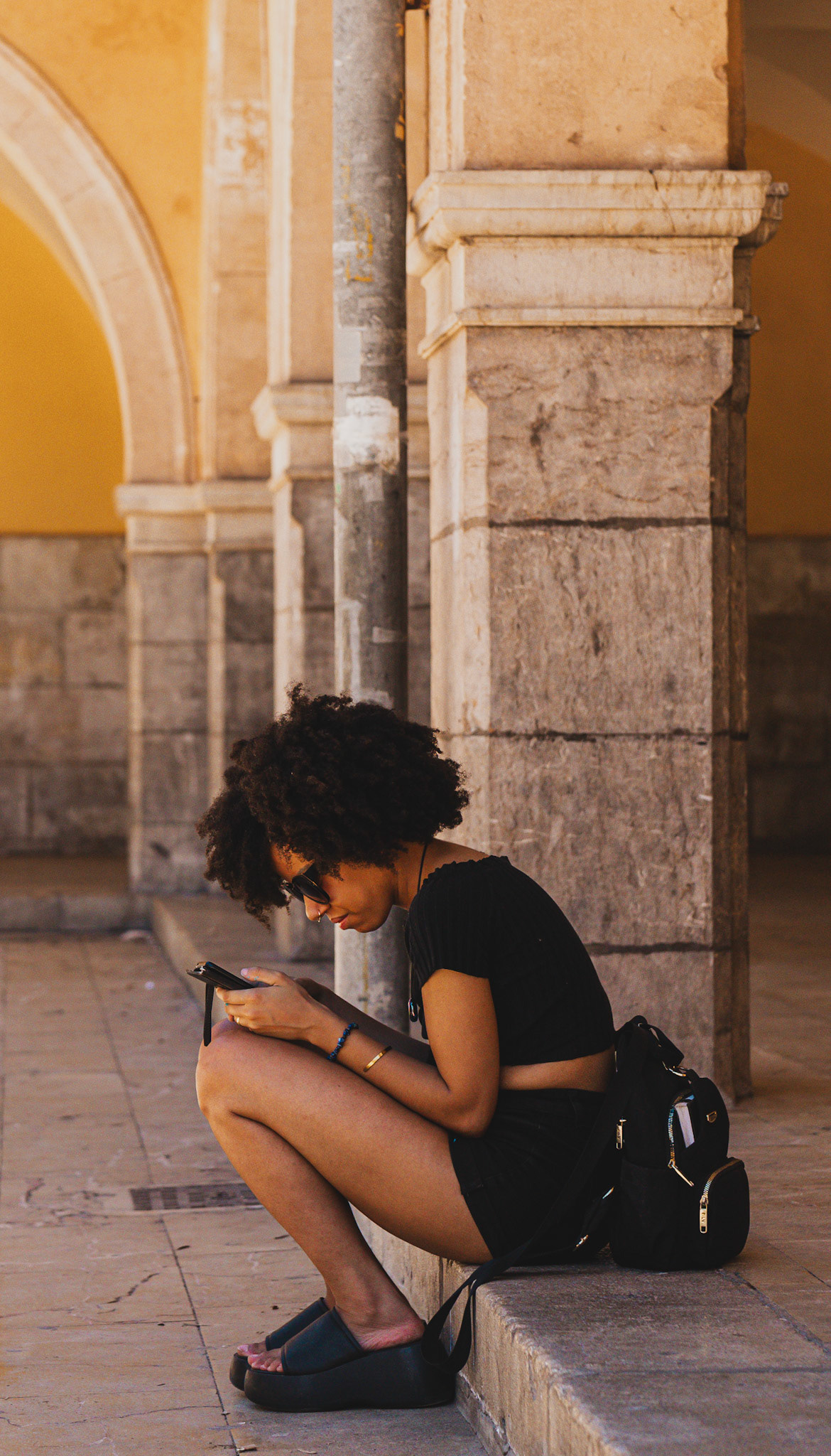 captured in front of mercat de l'olivar in palma, this photograph depicts a young woman engrossed in her phone, seated against the backdrop of historic stone arches. her relaxed posture and focused expression contrast with the bustling market scene, creating a moment of calm amidst the urban environment. the warm tones and textures of the architecture add depth and context, highlighting the blend of modern life and historical settings.
