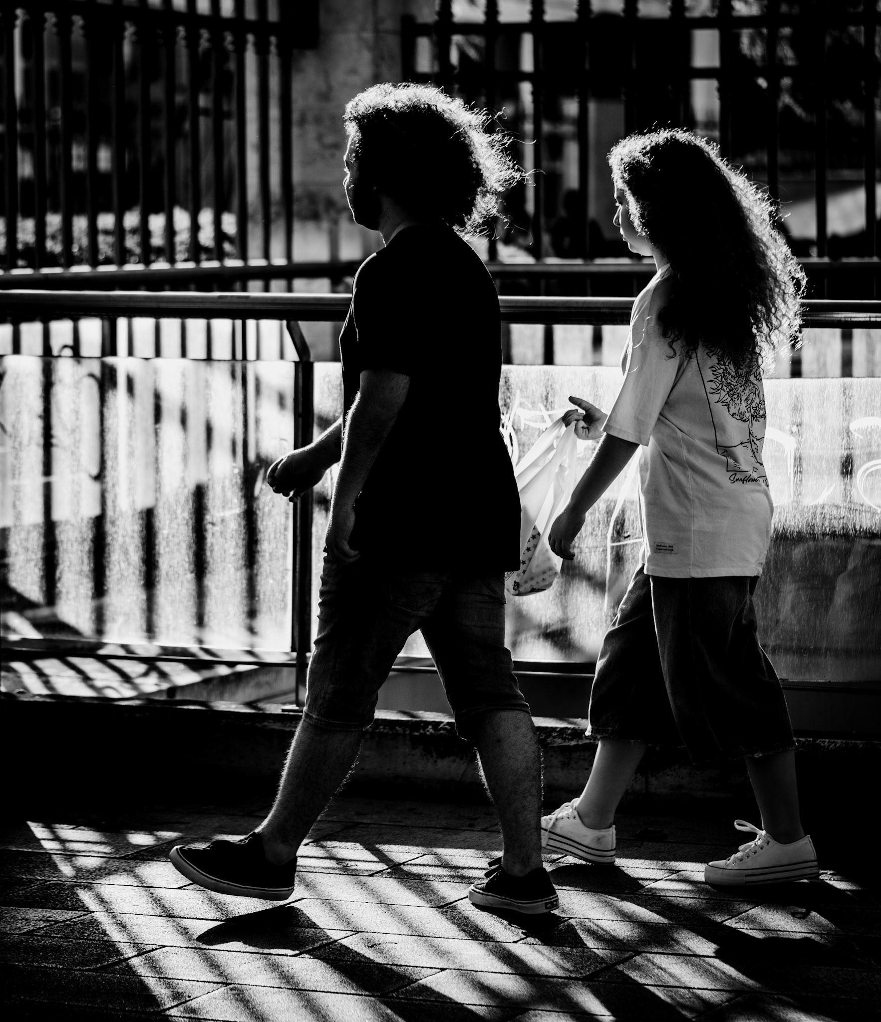 near plaça d'espanya, the interplay of lines, shadows, and the striking hair of two individuals creates a captivating scene. the dramatic backlighting highlights their silhouettes, making their curls glow with an ethereal quality. the crisscrossing shadows and architectural lines add a geometric rhythm to the composition, enhancing the visual intrigue of this moment.