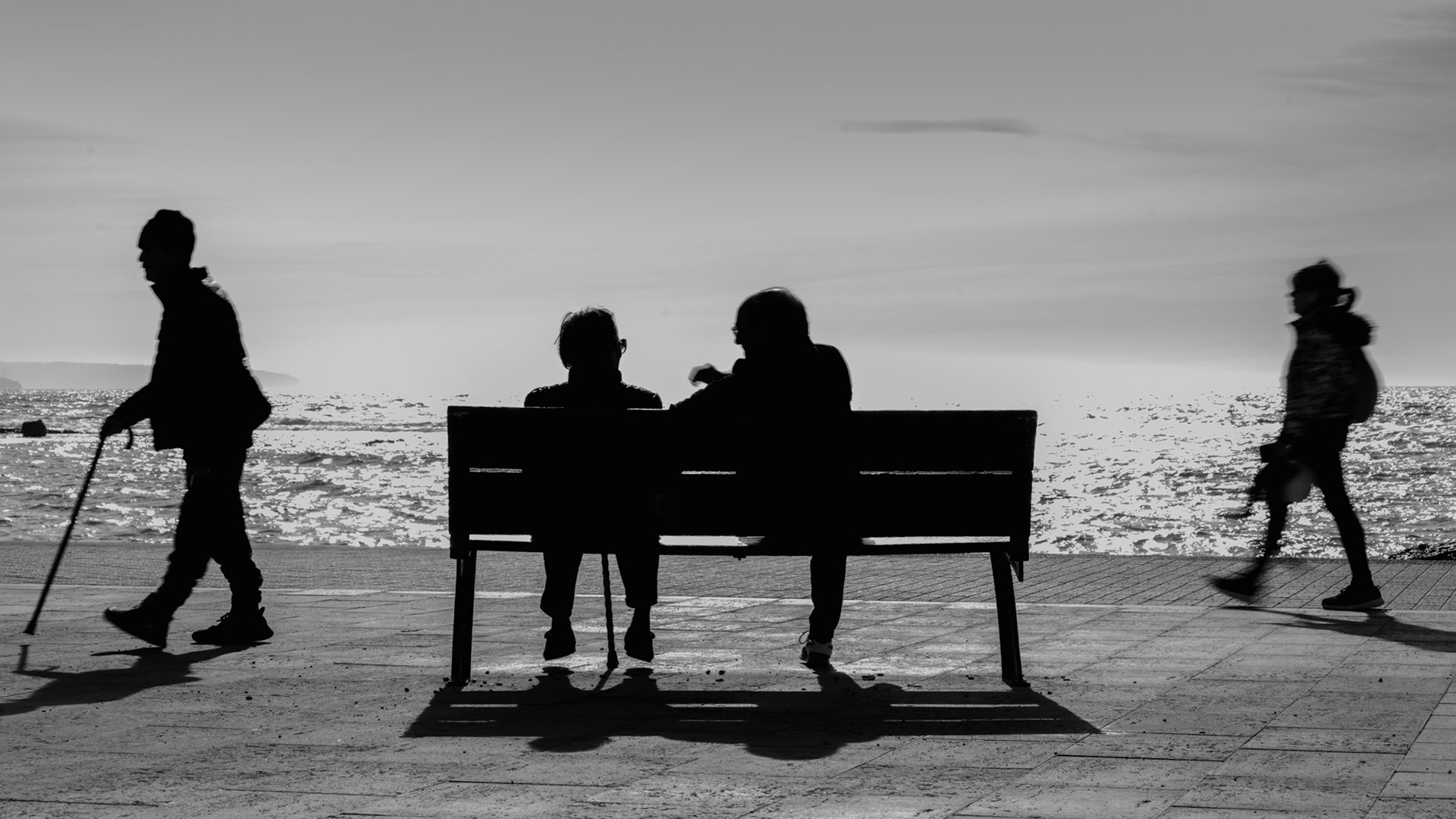 in the heart of portixol, where the sea whispers to the shore, a moment of tranquil conversation unfolds. the elderly couple, silhouetted against the shimmering sea, share silent stories as the sun casts gentle shadows at their feet. life moves around them – a man with a cane and a child with a teddy bear, embodying the passage of time. the bright horizon merges sea and sky, blurring the lines between reality and reflection. in this scene, stillness and motion dance together, painting a portrait of life’s ebb and flow.