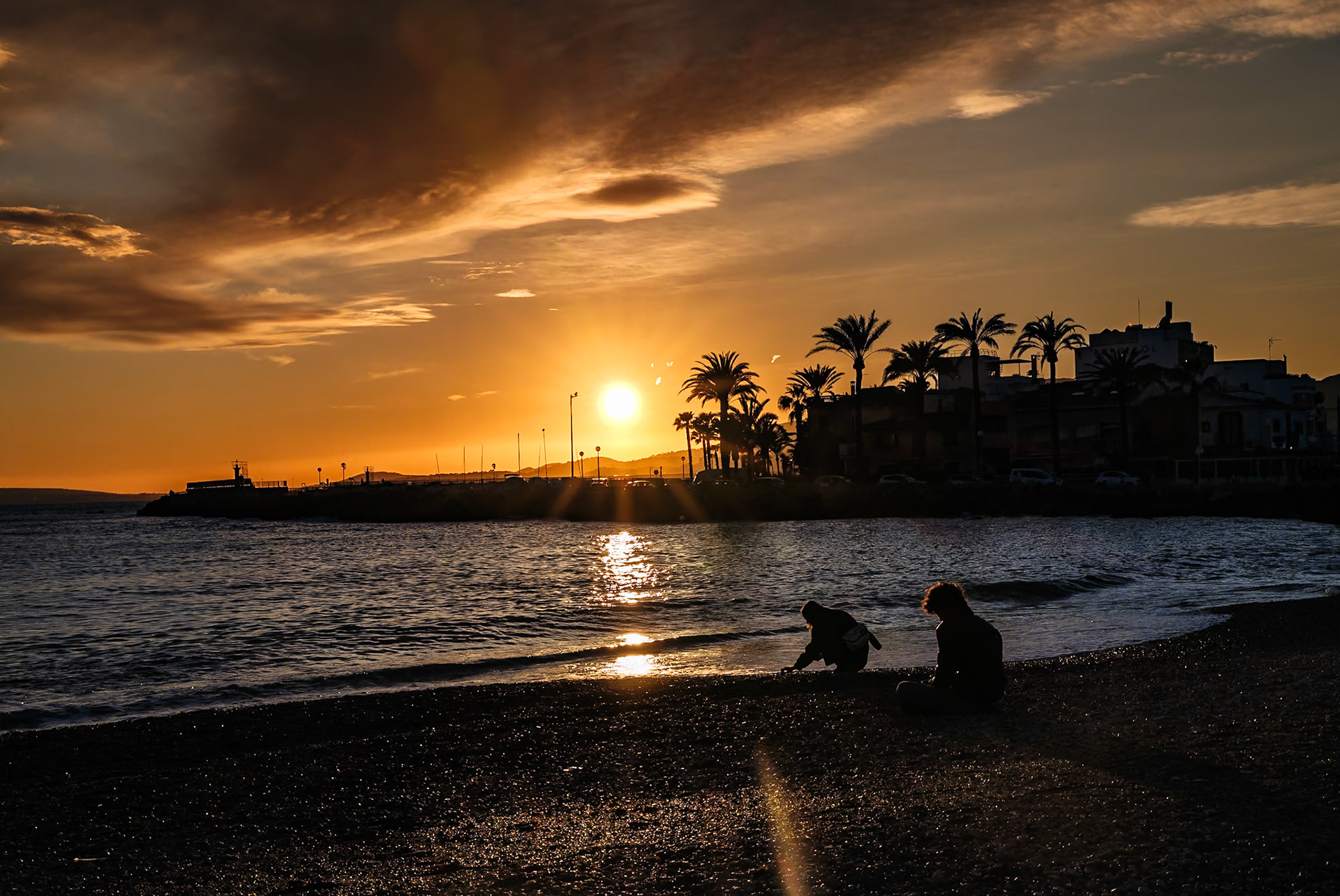 as the curtain of day descends, the shore of portixol transforms into a scene where the last act of sunlight unfurls its warm departure. not just a sunset is captured here, but a tender dialogue between the dimming daystar and the awaiting dusk. the sun, in its orb of flame, lingers close to the edge of the world, hesitant to conclude the daylight saga. its beams stretch over the undulating sea, painting the waters with strokes of molten gold and copper. on the shingle, pebbles shimmer, kissed by the day's final glance. figures are sketched against the backdrop, their outlines a part of the shoreline's ephemeral art. one stoops, perhaps collecting pebbles or the day's last moments, while the other sits, a solitary observer of the calm performance. here, in this frame, is a harmonious interplay of motion and stillness, of radiance and silhouette, all gathered to tenderly dismiss the daylight on shores where every twilight seems like a whisper of adieu.