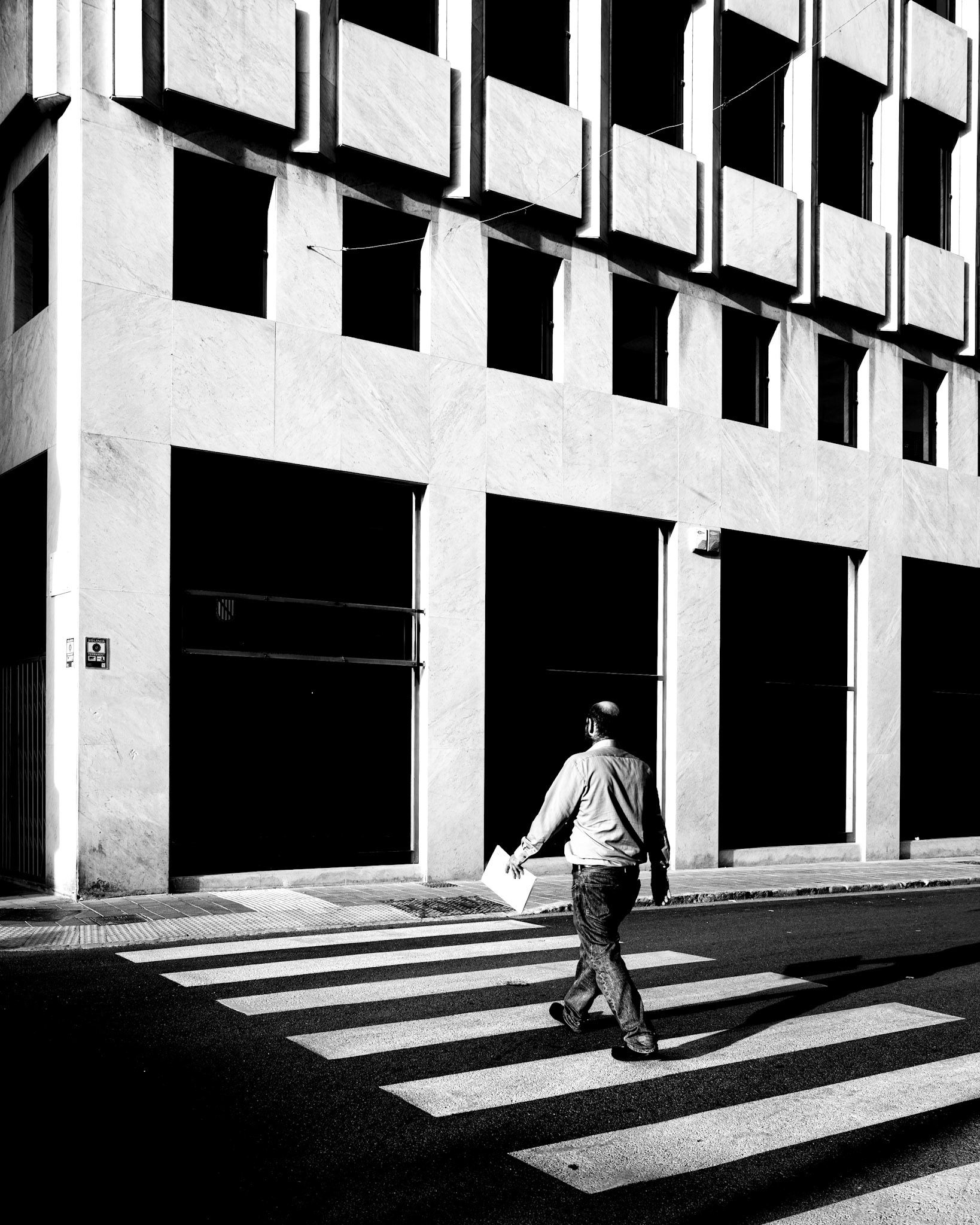 captured just around the corner from plaça espanya in palma, this image plays with geometry, contrast, and motion. the bold, repetitive patterns of the building’s facade create a striking architectural backdrop, while the zebra crossing guides the viewer’s eye to the man in motion. his casual stride and the paper in his hand add a human element to the rigid symmetry of the scene. the deep shadows and strong light evoke the sharp, sunny afternoons of mallorca, blending minimalism and street photography into a moment that feels both dynamic and timeless.