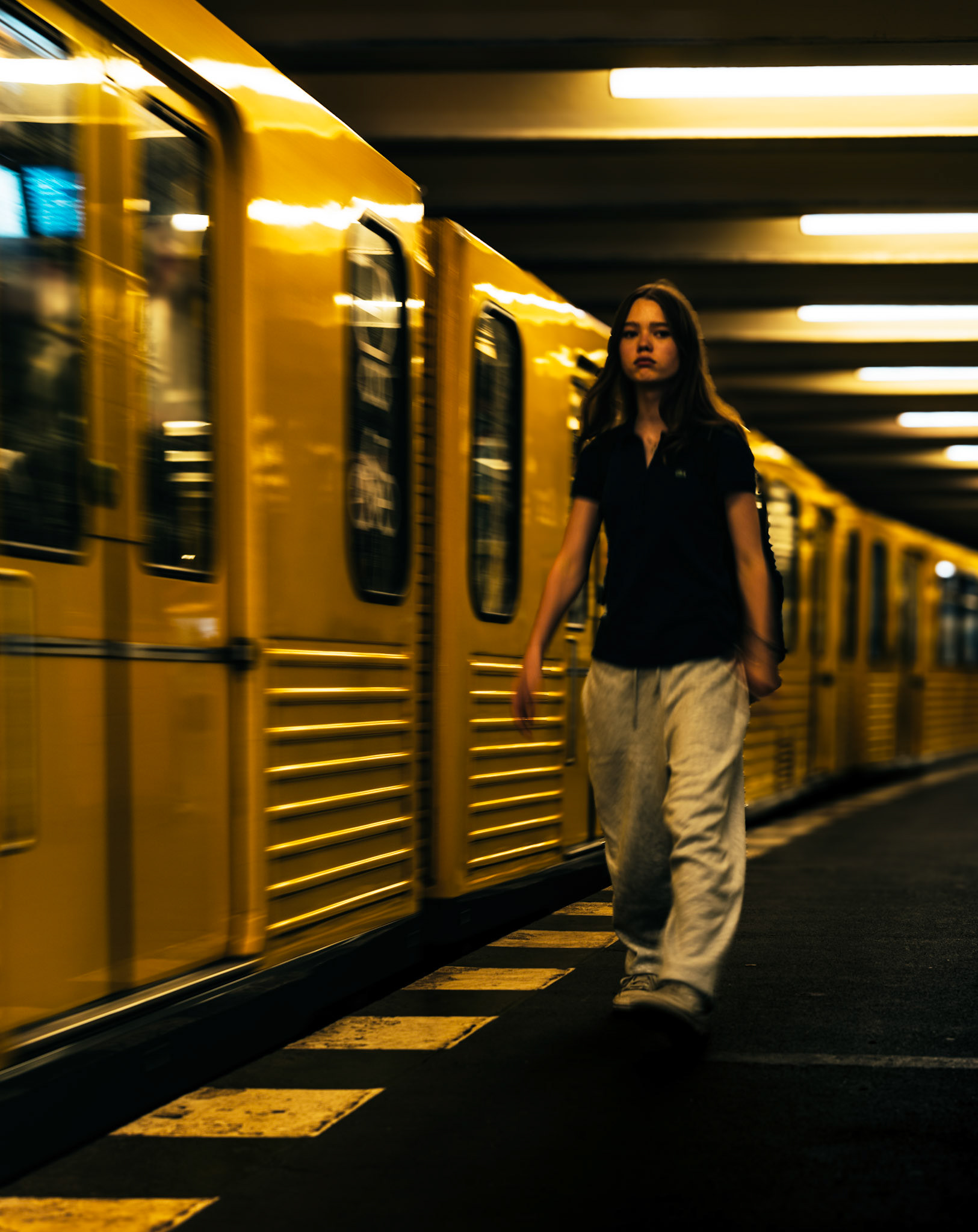 captured at the hausvogteiplatz u-bahn station in the heart of berlin mitte, this image speaks to the rhythm of city life, where moments are fleeting, and the lines between movement and stillness blur. the yellow train, iconic to berlin, streaks across the frame, a symbol of constant motion in an urban environment. yet, amidst this rush, the young woman walks with a purposeful calm, embodying the quiet resilience often found in the city's commuters. the cool tones of her attire contrast against the warm yellow of the train, creating a visual dialogue between the individual and the collective, the transient and the permanent.