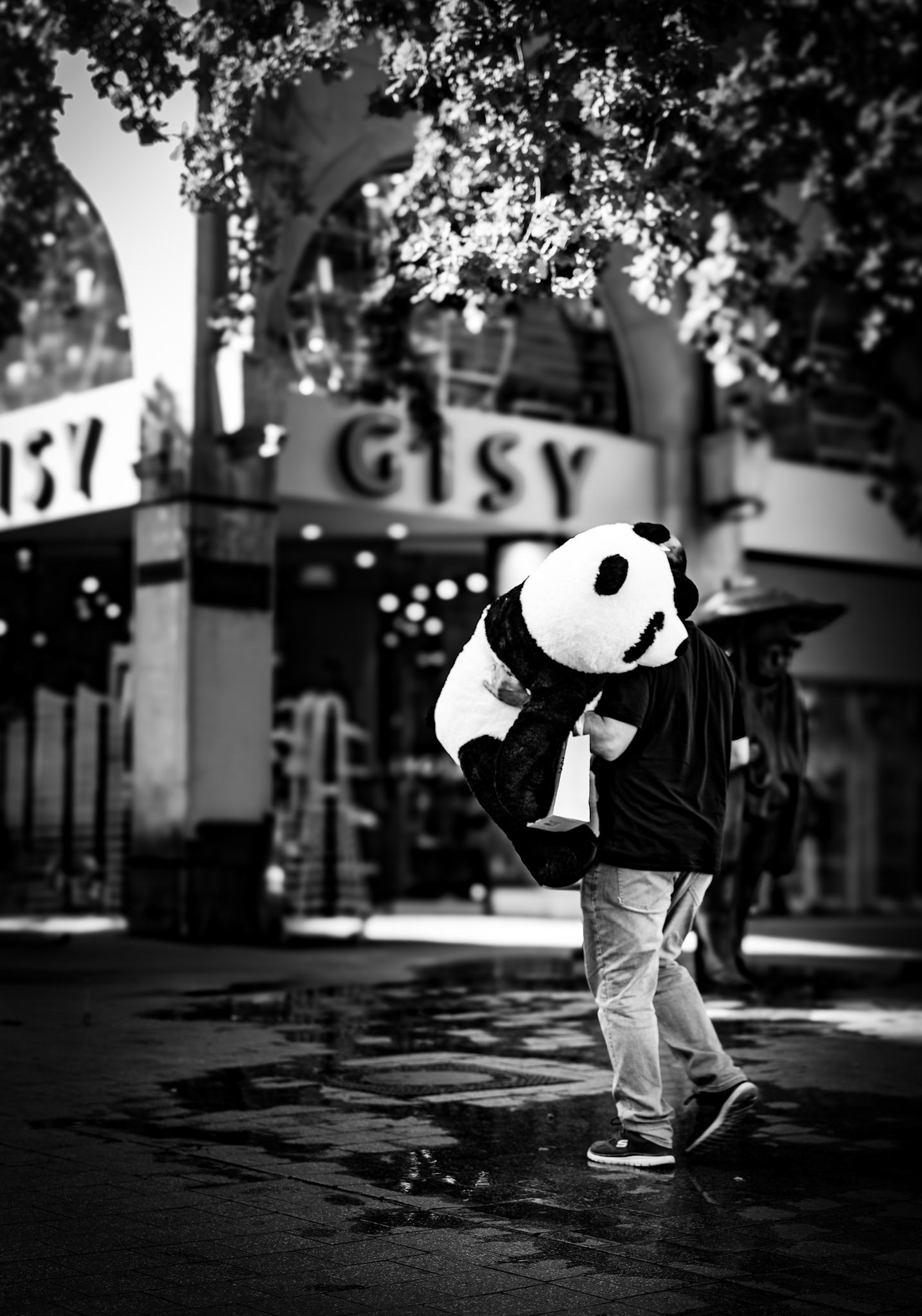 captured in hannover, not far from kröpcke, this photo freezes an unexpected city moment—a man carrying an oversized panda plush through the streets. the high-contrast black and white enhances the humor and absurdity of the scene, where the playful subject blends effortlessly into the urban backdrop. with his determined stride, the man almost becomes the hero of his own adventure, as if he's on a mission to bring the panda safely home. the reflection in the wet pavement adds another layer of reality, merging the ordinary with the whimsical. it's a moment where the mundane turns magical, and the city reveals its small, delightful stories.