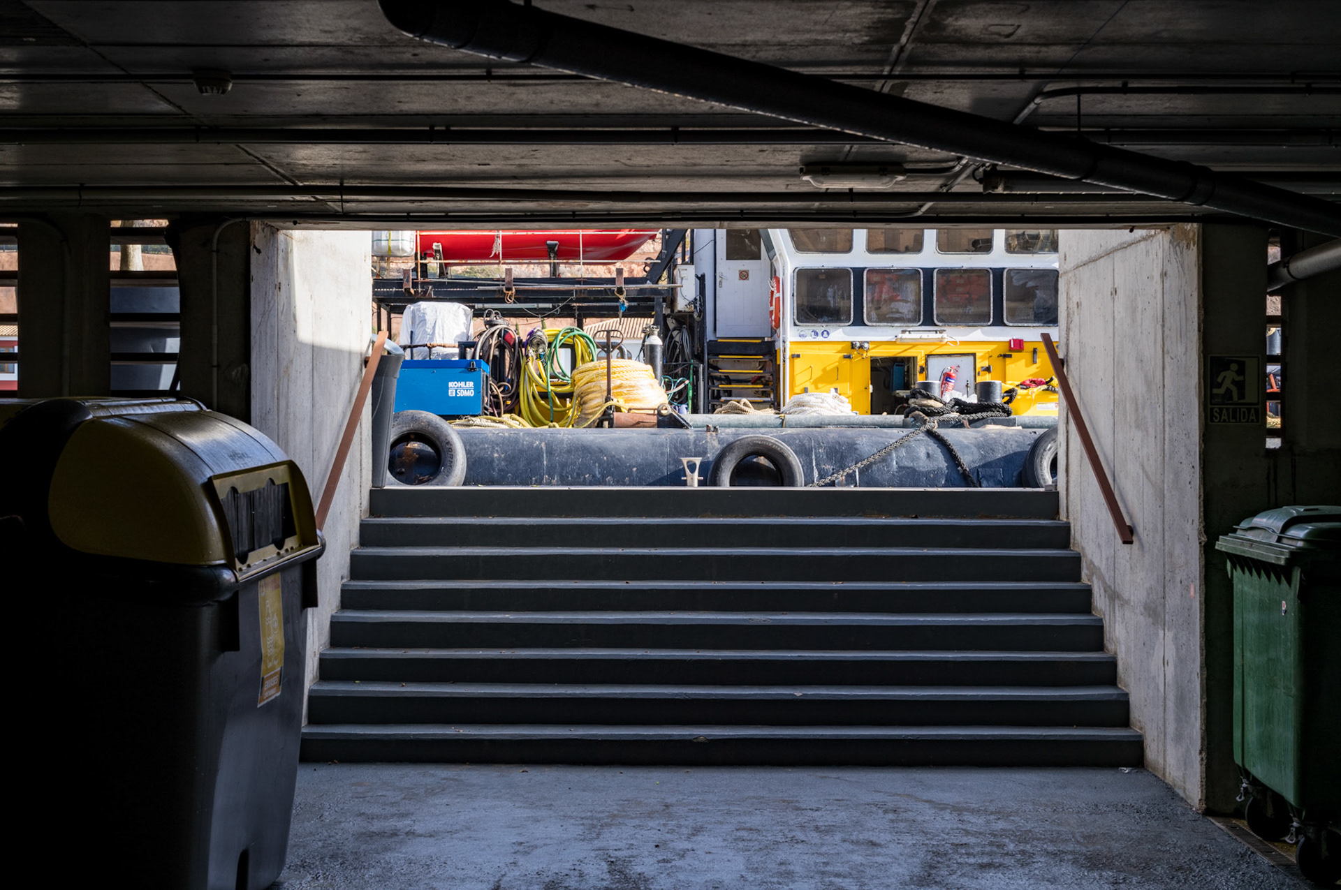 port adriano has been designed by philippe starck and is a relatively new harbour in the south-west of mallorca, between santa ponca and magaluf. apparently they seem to have some works so when i was leaving the underground parking i enjoyed the view to this industrial vessel. "This photograph depicts an urban scene viewed from within a dimly lit garage or underground area, looking out towards a brightly lit outdoor space. In the foreground, there are grey concrete steps leading upwards, flanked by white walls. At the top of the stairs, there is a view of a busy construction or maintenance site, indicated by the presence of various equipment and vehicles. There are large reels of cable. The contrast between the shadowy interior and the illuminated exterior is striking, with the daylight revealing the details of the industrial equipment and machinery. It's a candid and unembellished snapshot of harbour life, blending elements of architecture, urban planning, and industrial activity."