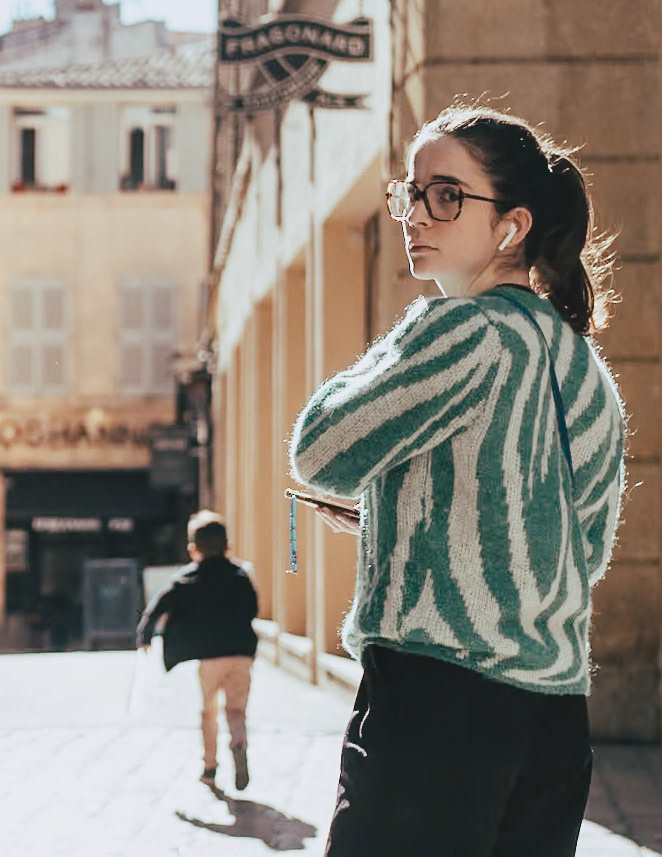captured in the historic streets of aix-en-provence, this image features a young woman pausing to look back, her expression a blend of thoughtfulness and casual awareness. dressed in a striking green striped sweater, she stands in contrast against the old-world charm of the sunlit cobblestone street. her modern style juxtaposes with the timeless architecture around her, creating a visual dialogue between the past and the present. the composition suggests a moment caught in transition, reflecting the vibrant life of the city as it moves around her. it's a snapshot of contemporary life set against the backdrop of a city rich with history, inviting viewers to ponder the layers of stories these streets hold.