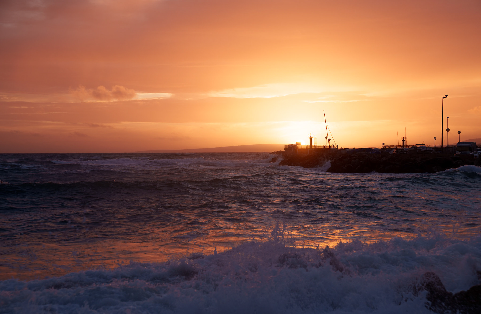 the photo captures the tranquil yet dynamic scenery of portixol, a charming seaside district of palma de mallorca. after stormy weather, the setting sun casts a warm orange glow over the turbulent ocean, illuminating the sky and waves, creating a serene yet powerful display of nature's beauty at dusk.