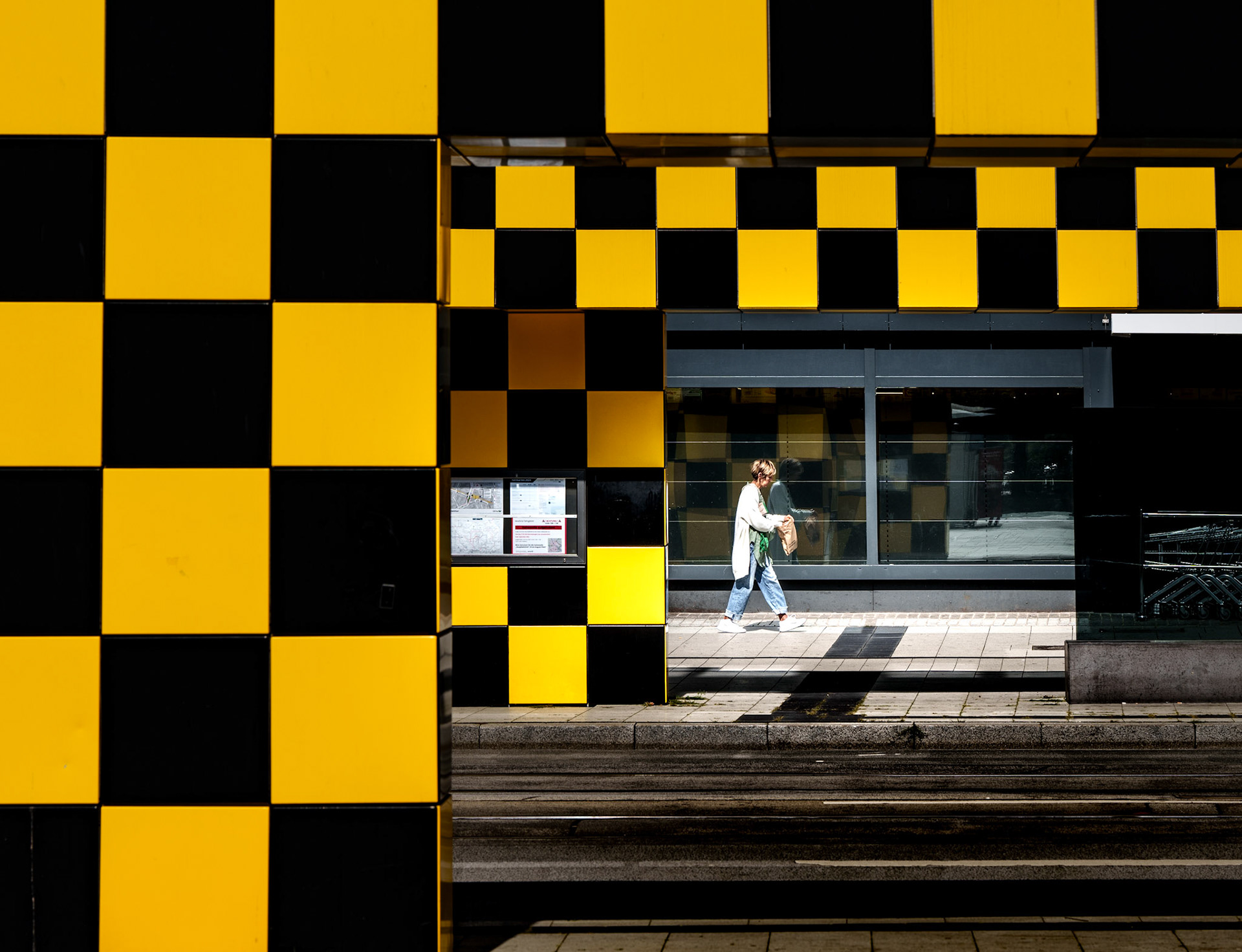 a moment frozen in the rhythm of the city—where geometry and life intersect. the stark black and yellow checkered facade, bold and unapologetic, forms a rigid grid that slices through the urban landscape. within this pattern, a lone figure moves with quiet determination, caught mid-stride in the sun's embrace. her shadow stretches out, echoing her path, as she walks through the space between the lines, an organic element amid the structured chaos. the play of light and shadow adds depth, while her presence brings warmth and humanity to the cold, calculated architecture around her. it’s a fleeting dance between order and freedom, stillness and motion, captured in a heartbeat.