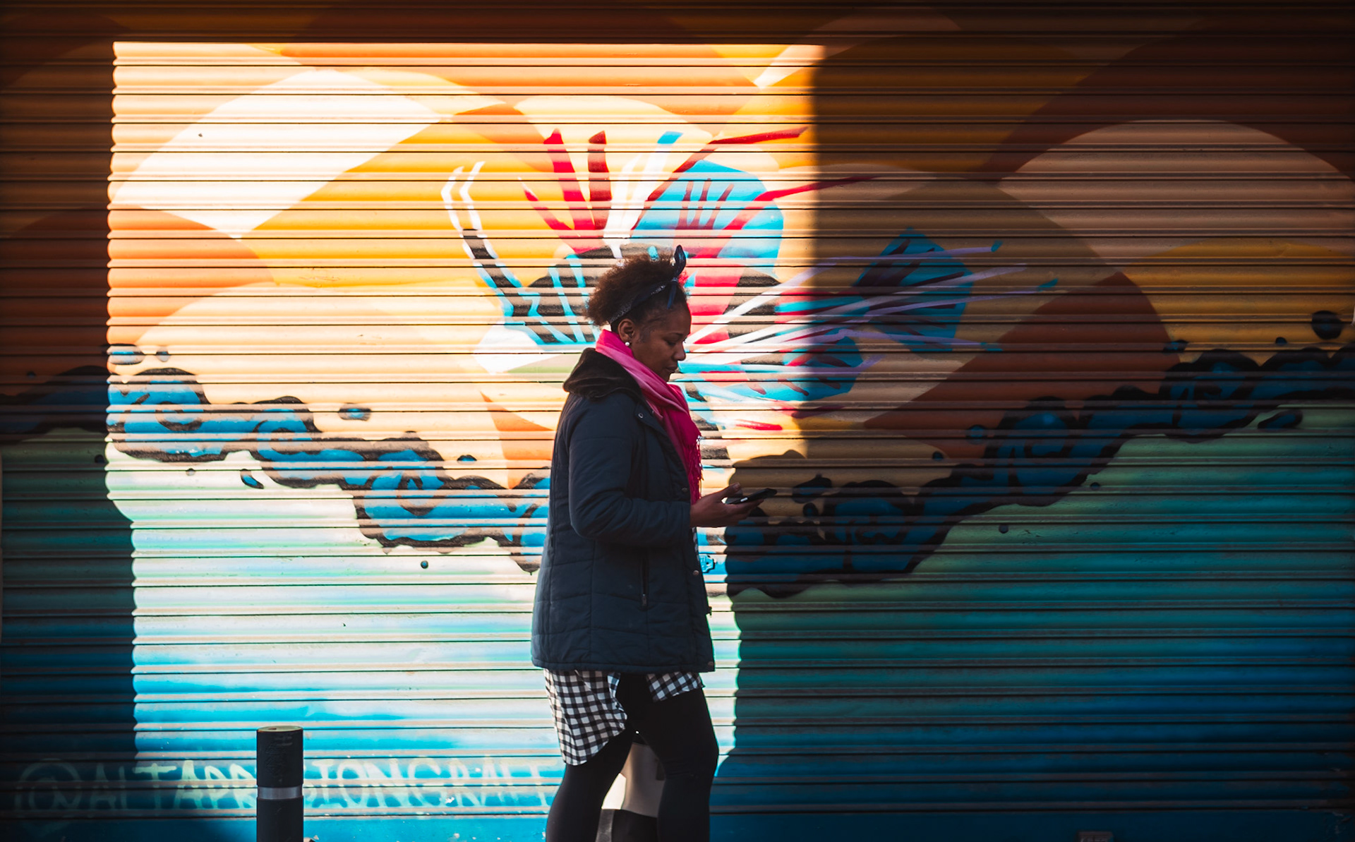 in the heart of palma's joan miró, the play of light cuts sharply through the urban canvas, revealing layers of color that dance vibrantly on a mundane morning. a woman, engrossed in the digital world, glides past a graffiti-splashed shutter that tells a more vivid tale of the street than the fleeting glances of passersby might suggest. her silhouette, softened by the backlight, juxtaposes with the spray-painted hues, crafting a narrative of movement amidst stillness. the shadows cast by the morning sun enhance the graffiti's bold lines and shapes, breathing life into the inanimate, and imprinting on her journey a fleeting touch of artistry.