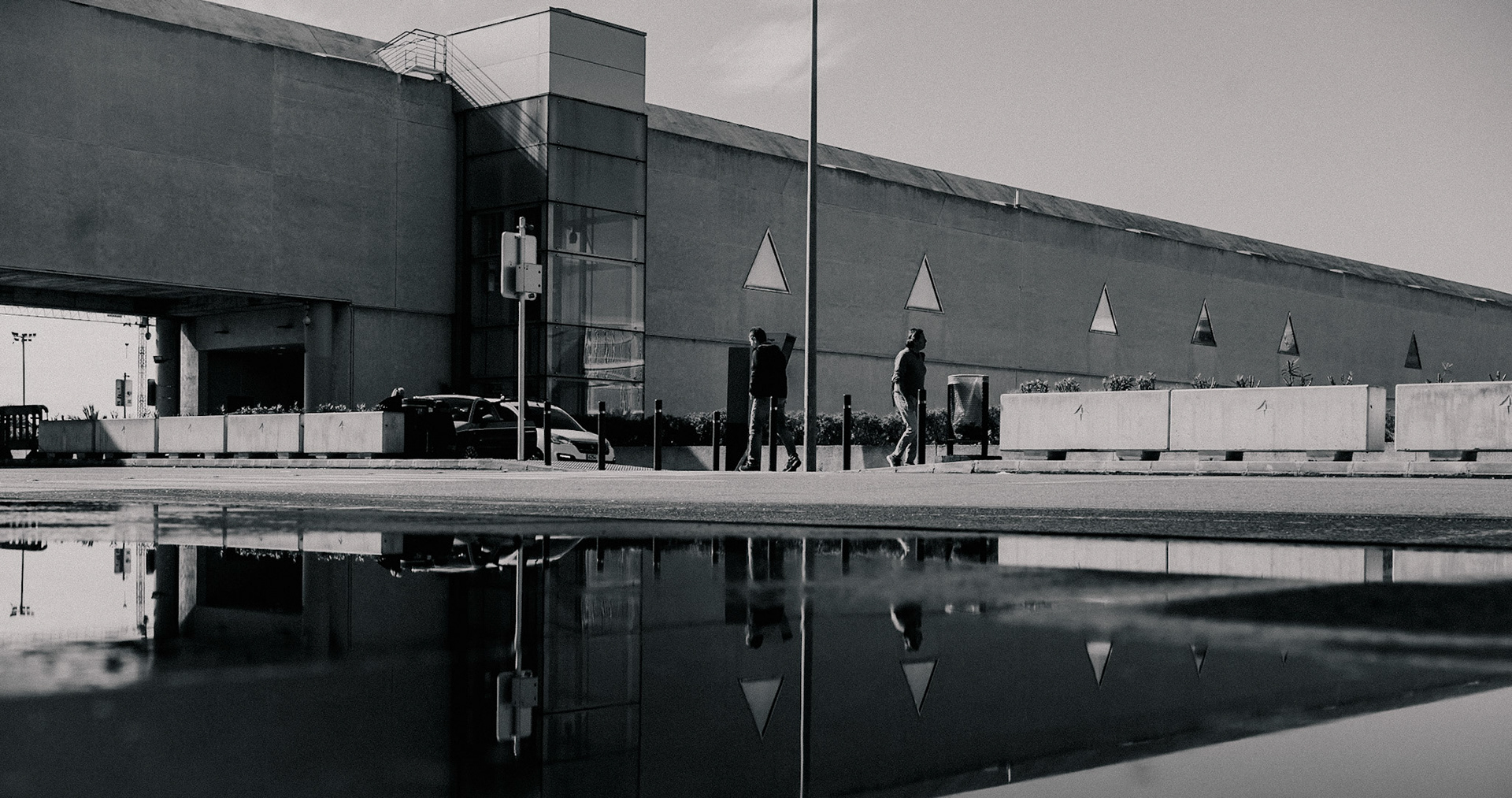 after the storm, the world turns reflective, quite literally. outside the bustling gateway of palma's airport, where farewells linger and reunions spark in quick succession, the heavy rain had left behind a mirror on the ground. it's a scene split between two realms – the upper world carries on, oblivious to its upside-down twin below. travelers walk past, reflected as specters in the transient pool, doubling their presence in this transient space. in the calm after the downpour, even the rigid lines of modern architecture seem to soften, to flow with the whims of water and light. a passerby stops mid-step, perhaps caught in the realization that every journey leaves a ripple, seen or unseen.