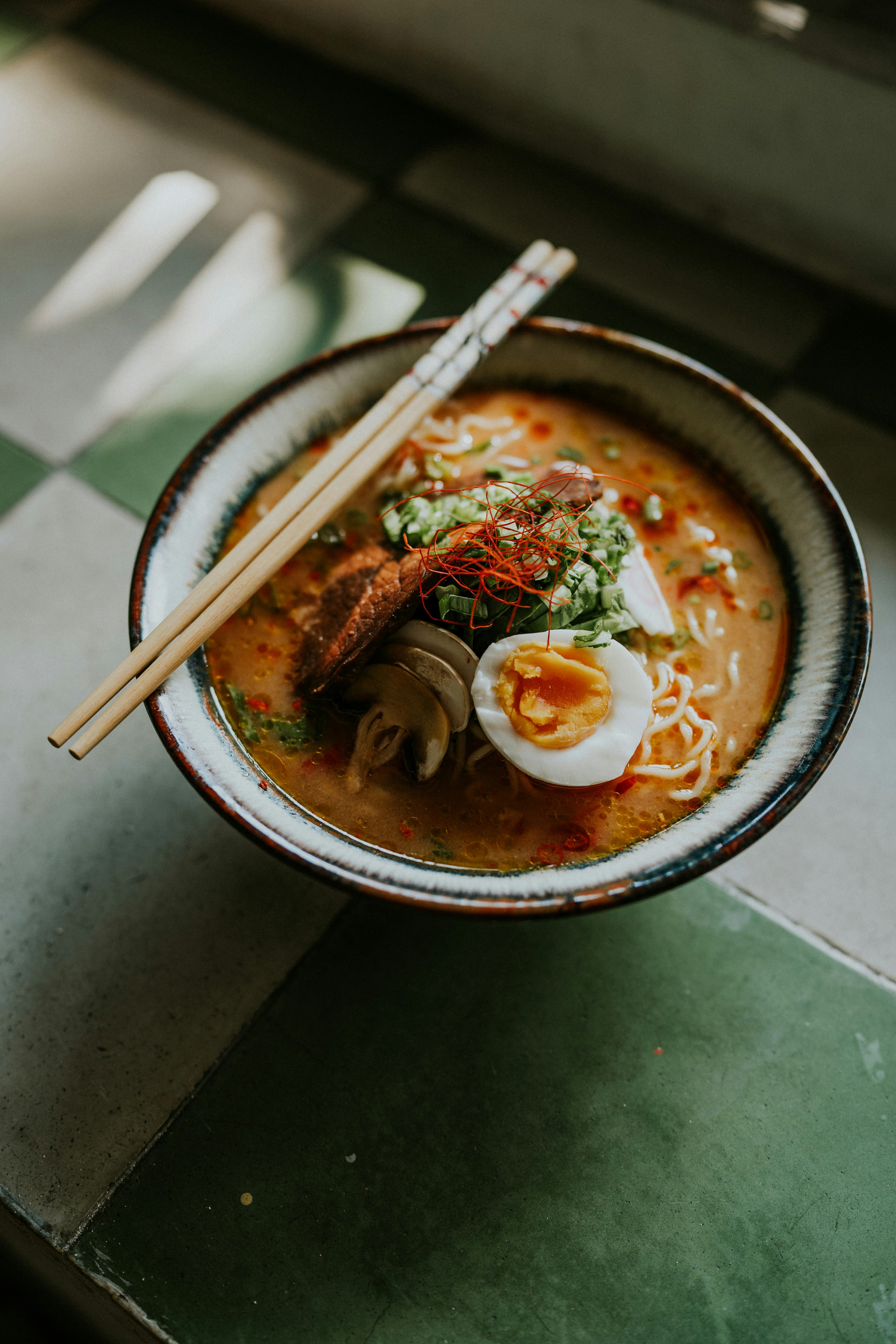 Image of a hot ramen served on a bowl with chopsticks ready to eat. Topping  includes noodles, boiled egg, mushrooms, and beef.